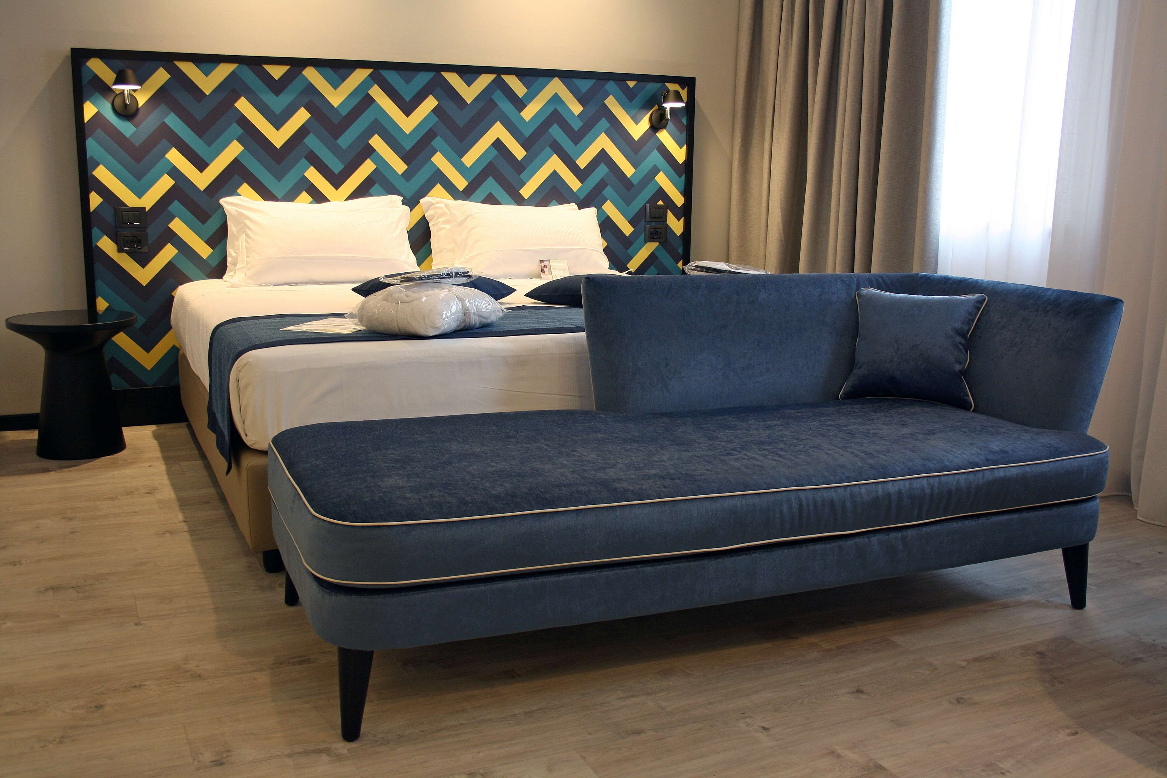 Ett modernt hotellrum med en säng, en blå soffa och en mönstrad sänggavel.