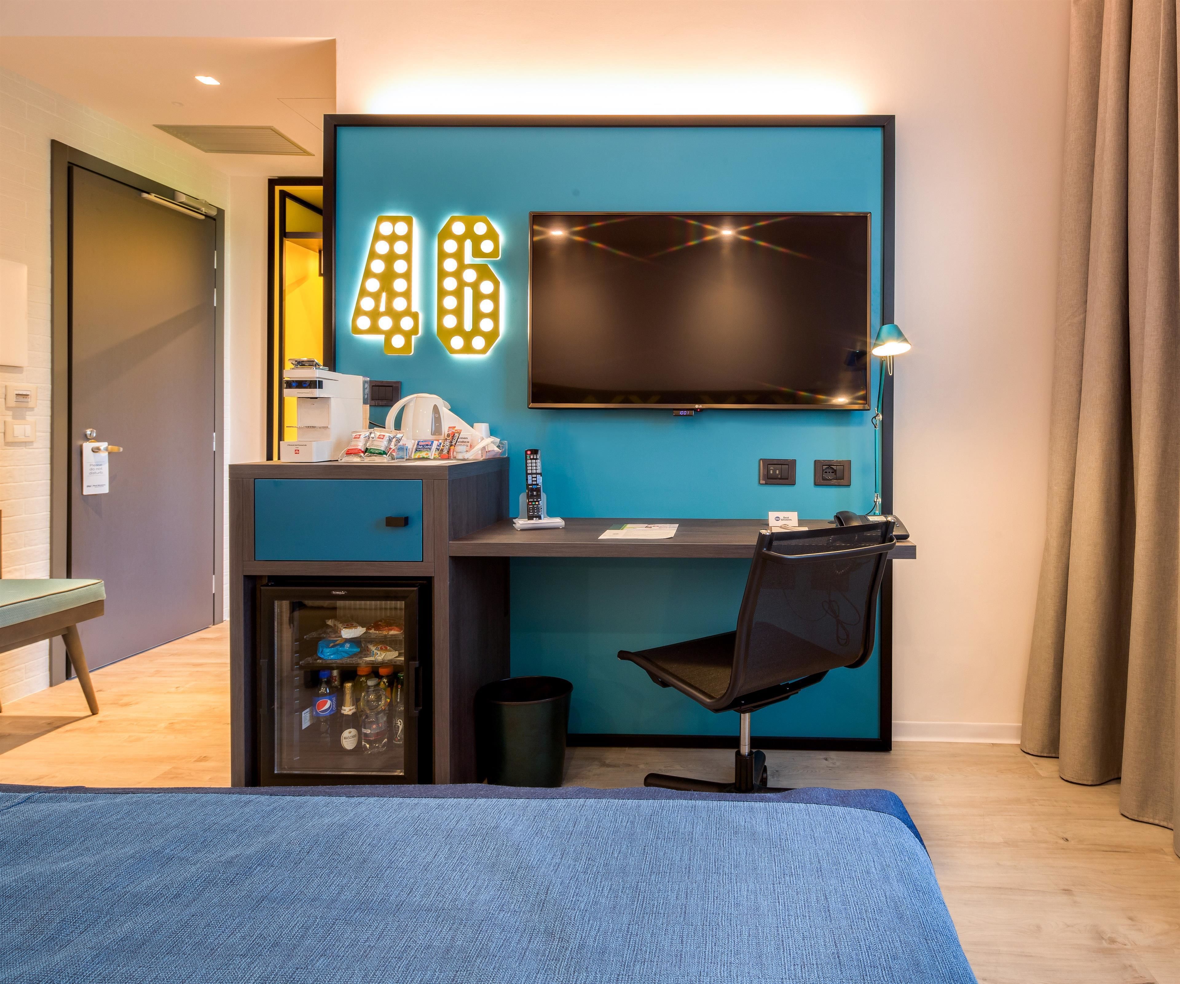Ein modernes Hotelzimmer mit blauer Akzentwand, einem Schreibtisch mit Computer, Fernseher und Minibar.