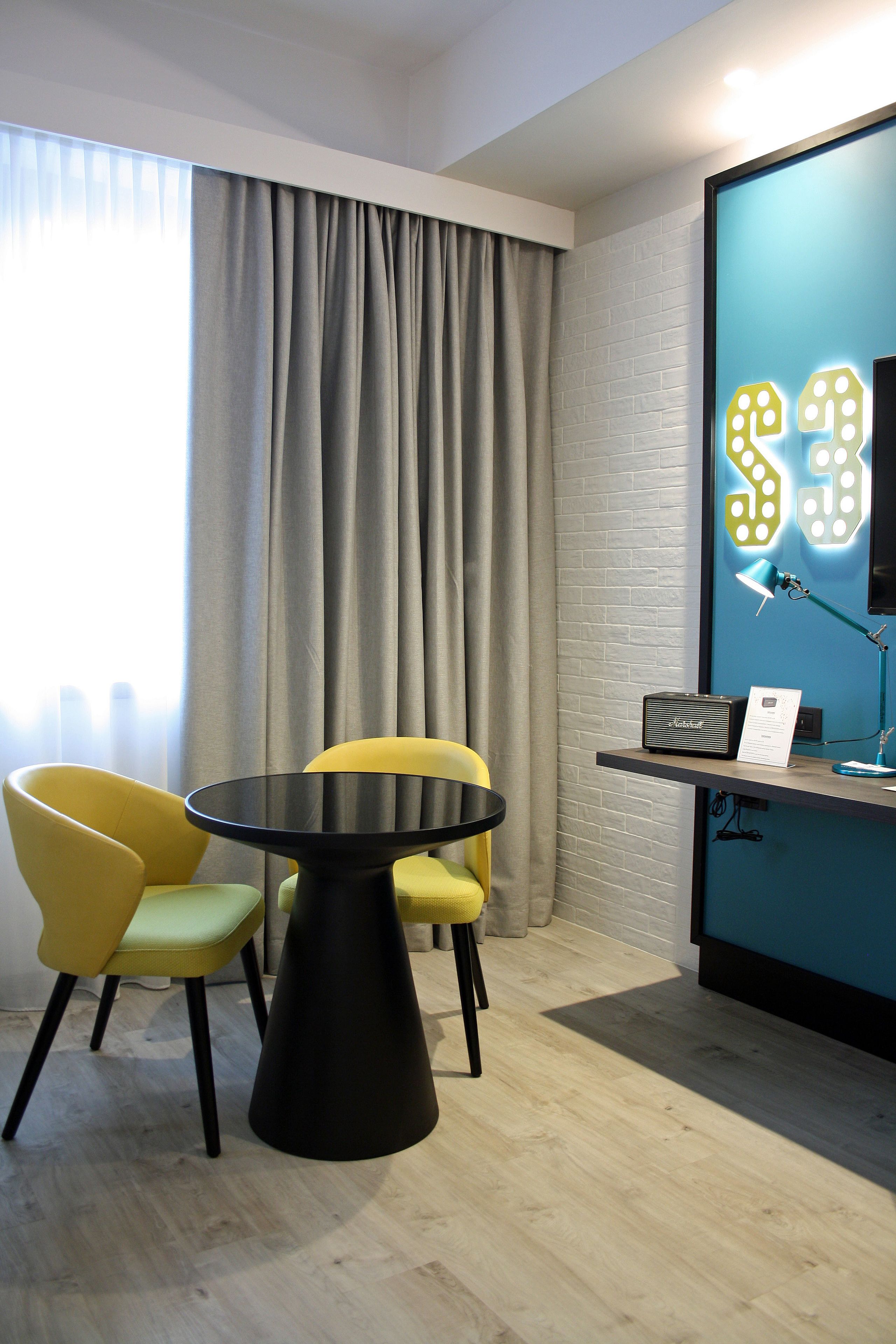 A modern room with a round black table, two yellow chairs, a grey curtain, a blue wall with a light-up sign, and a desk with a lamp and a speaker.