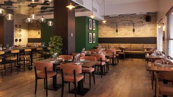 A restaurant with multiple dining tables and chairs, a green accent wall, and hanging pendant lights.