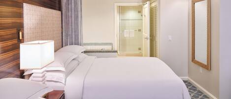 A hotel room with a large bed, a bedside table with a lamp, a mirror, and a shower area visible through an open door.