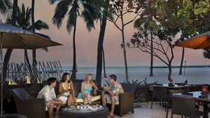 A group of people seated around a round table with drinks, surrounded by wicker furniture and umbrellas, overlooking a beach with palm trees and a sunset. - Hilton Grand Vacations Club Grand Waikikian Honolulu (Honolulu)