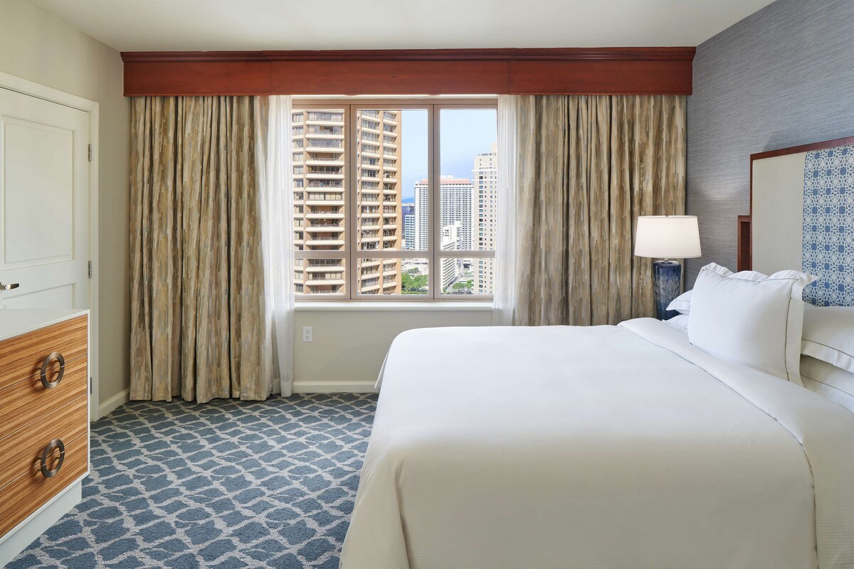 Ein Hotelzimmer mit einem großen Bett, einem Schminktisch, einem Fenster mit Vorhängen und Blick auf hohe Gebäude.
