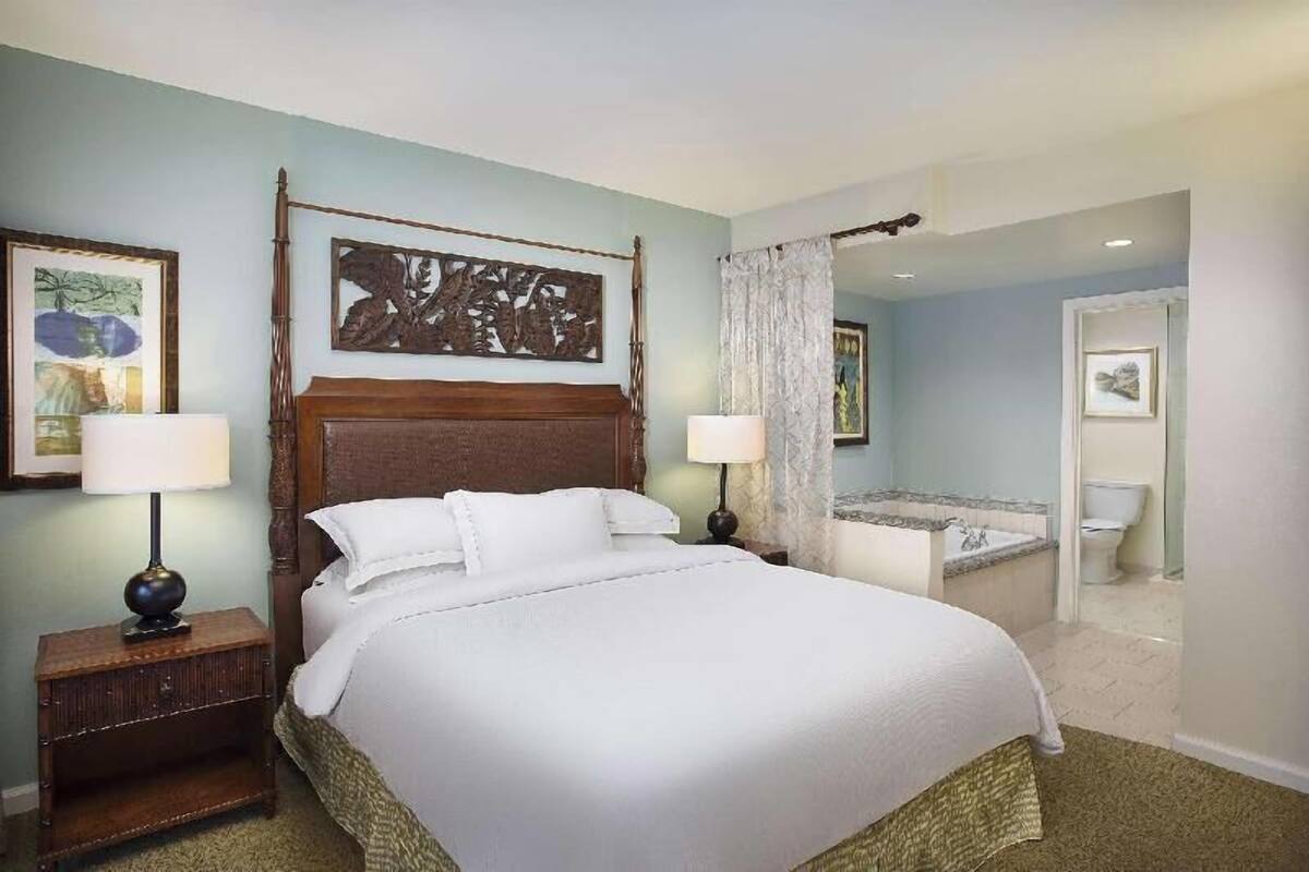 Une chambre à coucher avec un grand lit, deux tables de chevet avec des lampes, une tête de lit et une salle de bain avec une baignoire et des toilettes visibles à travers une porte ouverte.