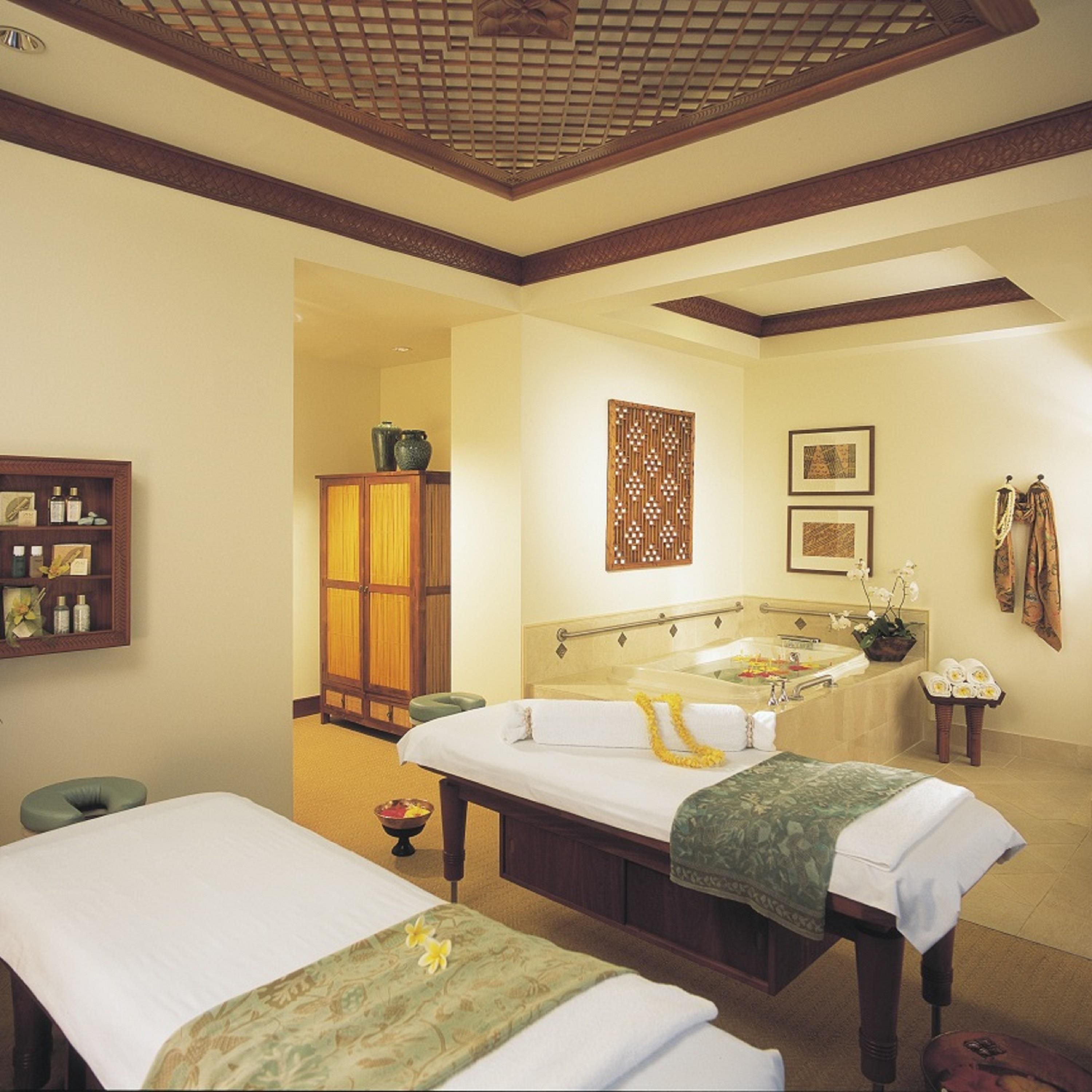 A spa room with two massage tables, a bathtub, and a wooden cabinet with bottles.