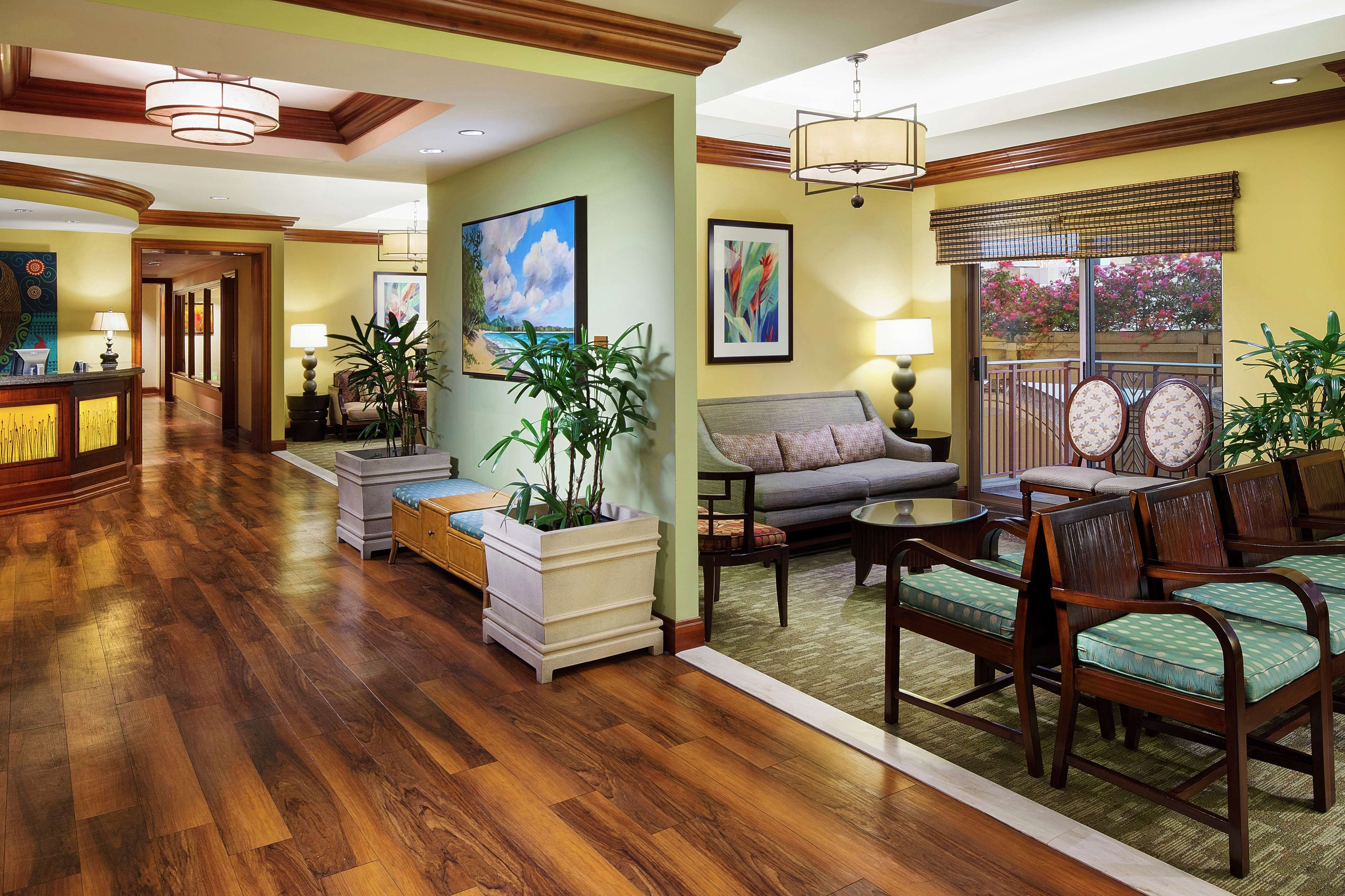 A spacious living area with hardwood flooring, a sofa, a coffee table, and a dining area with chairs. There are potted plants and artwork on the walls.