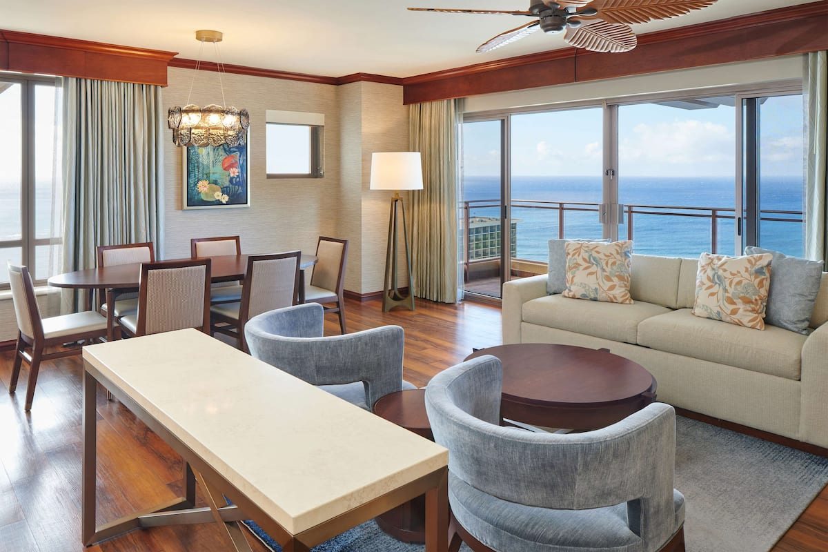 Een hotelkamer met een eettafel, stoelen, een bank en een groot raam met uitzicht op de oceaan.