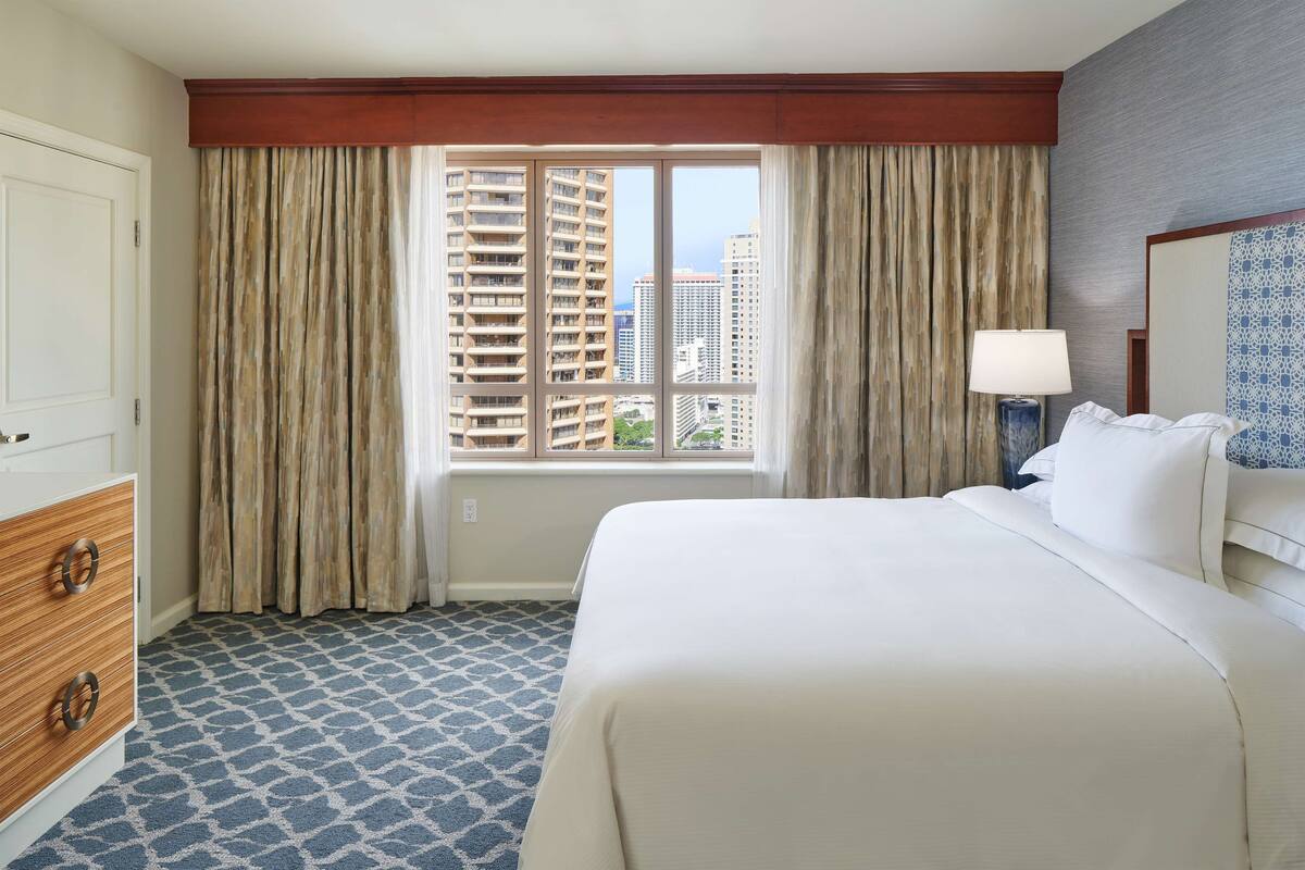 Een hotelkamer met een groot bed, een dressoir, een raam met gordijnen en uitzicht op hoge gebouwen.