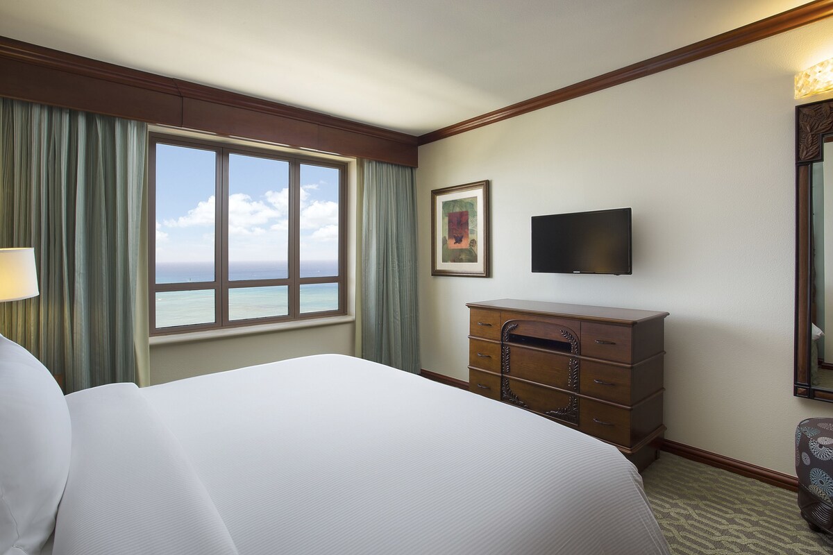 Habitación de hotel con una cama grande, un tocador, un televisor y una ventana con vistas al océano.