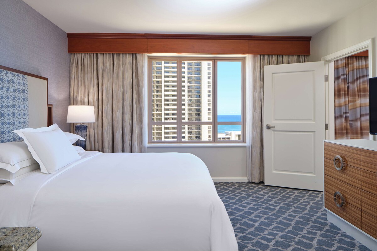 Une chambre d’hôtel avec un grand lit, une commode, une fenêtre donnant sur des immeubles et une porte.