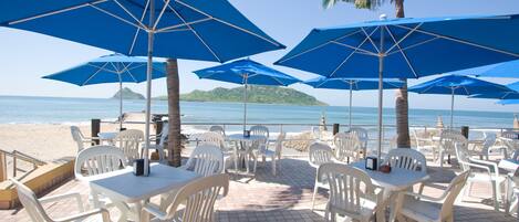 A beachside restaurant with outdoor seating, blue umbrellas, and white plastic chairs.