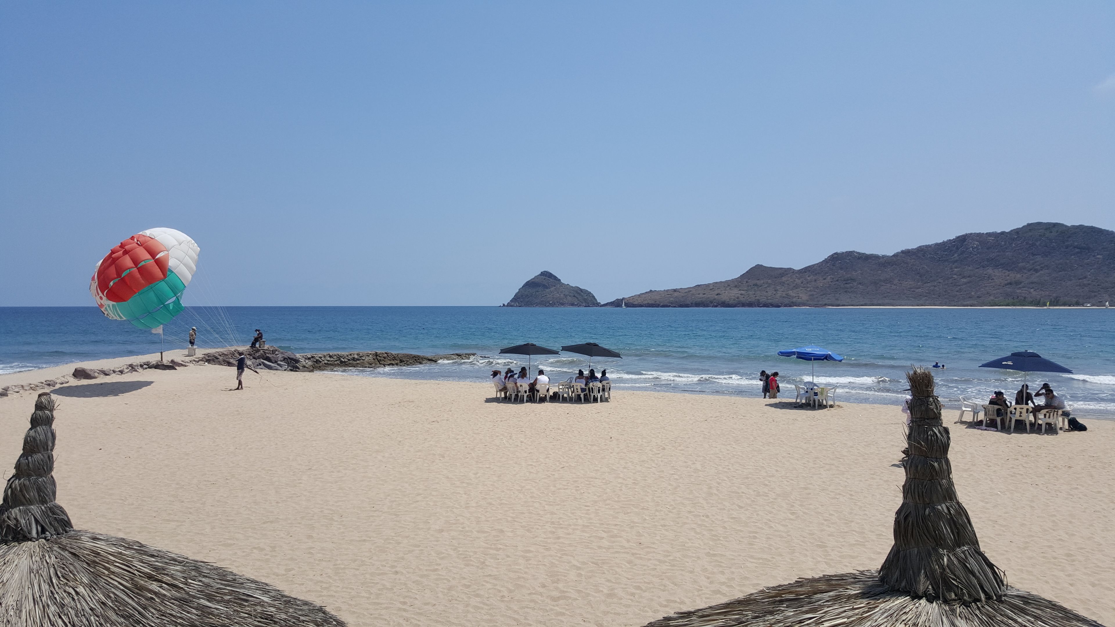 A beach with a parasail, people sitting under umbrellas, and rocky outcrops.