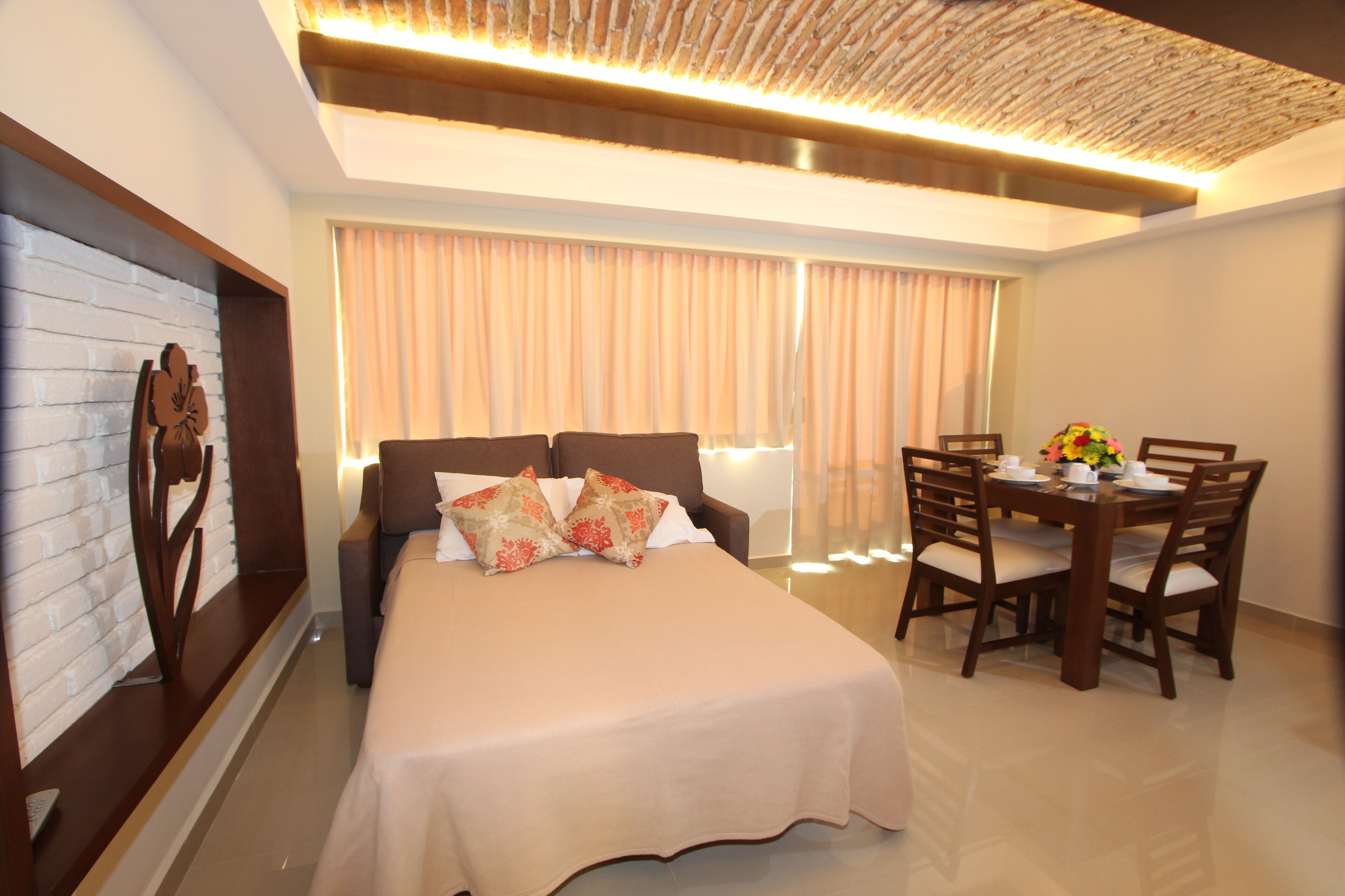 A modern hotel room with a large bed, a dining table with chairs, a decorative wall, and a large window with curtains.