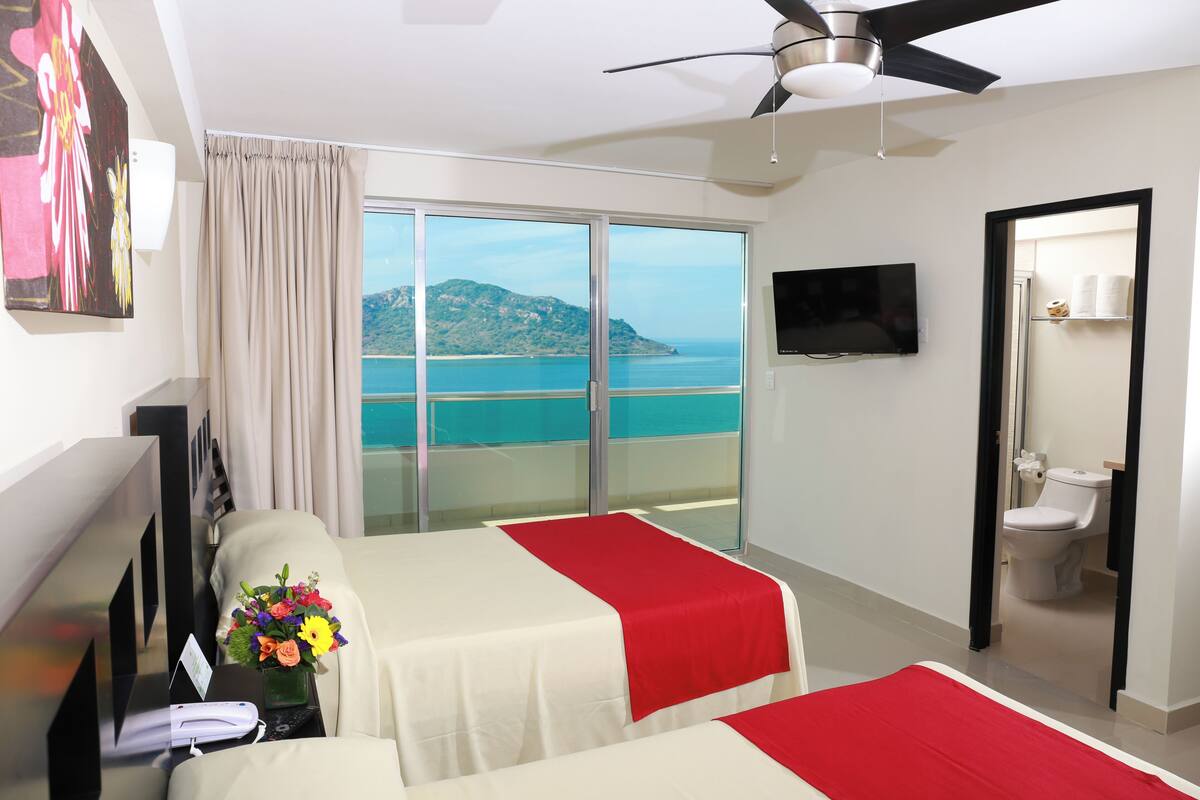 Ein Hotelzimmer mit zwei Betten, einem Fernseher, einem Schreibtisch mit Telefon und Blumen, einem Balkon mit Meerblick und einem Deckenventilator.