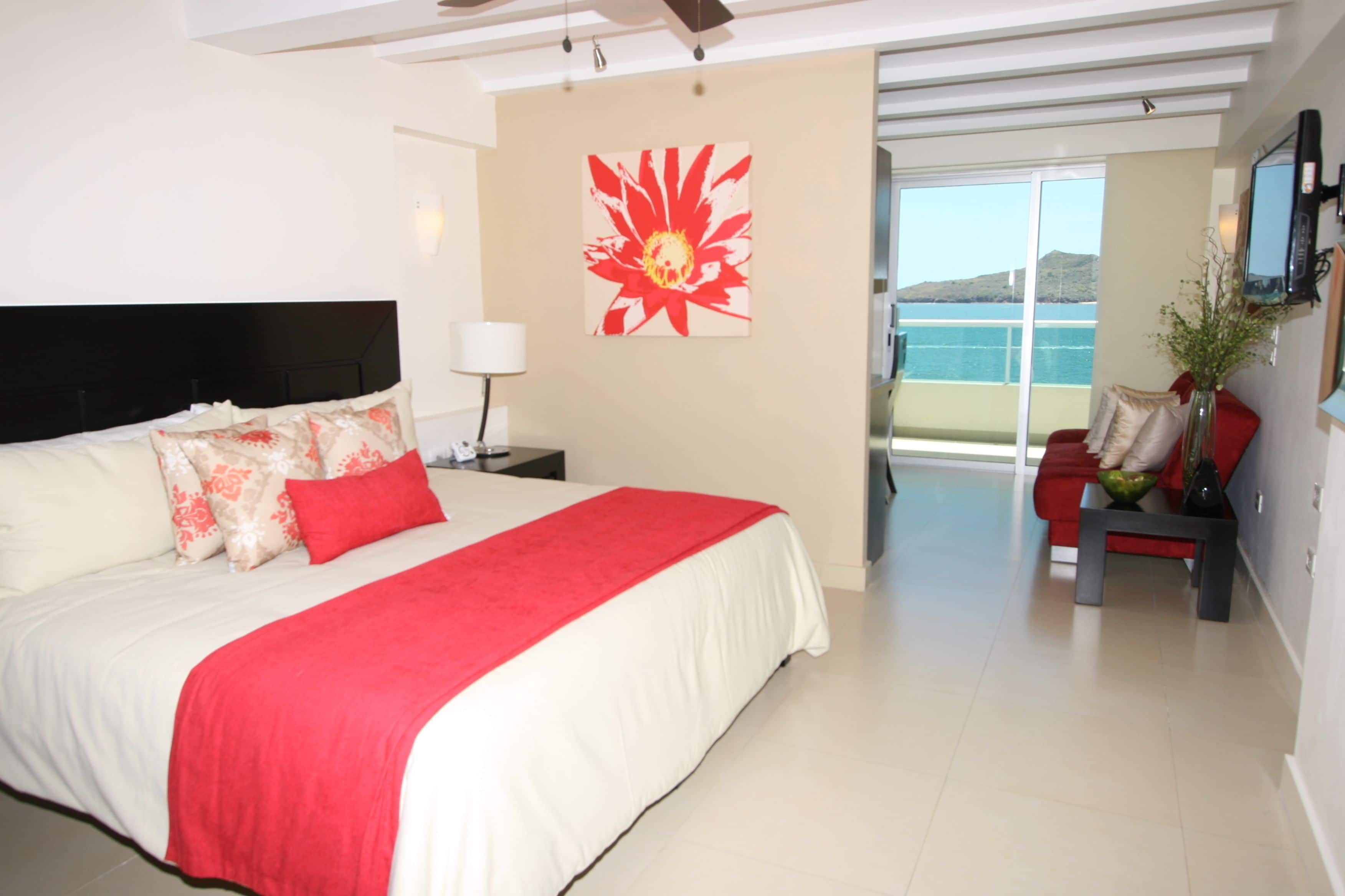 A bedroom with a large bed, a red and white bedspread, a red pillow, a nightstand with a lamp, a red chair, a vase with flowers, and a large window with a view of the sea.