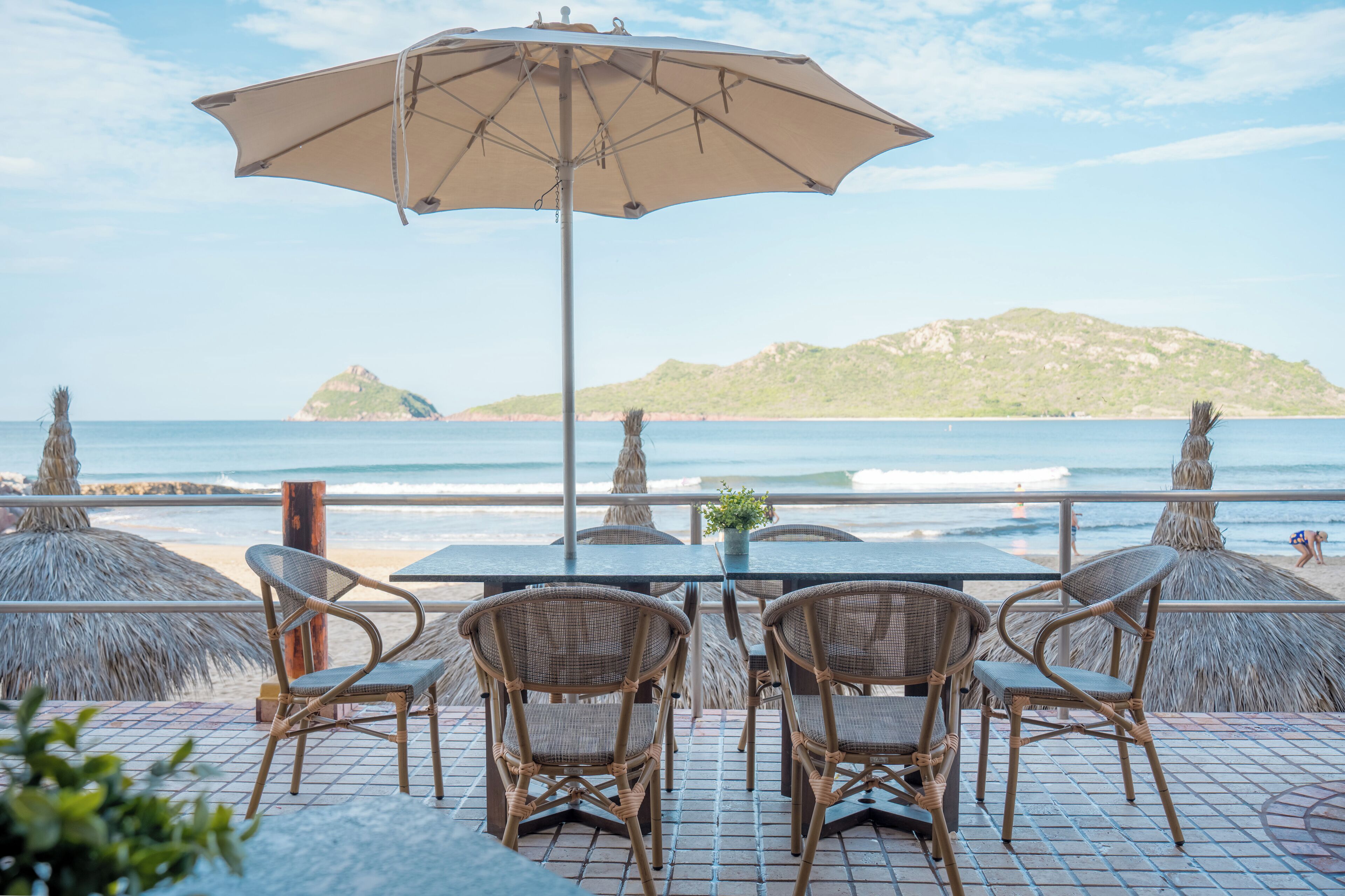 A table with four wicker chairs and a large umbrella, overlooking a beach with a view of the ocean and a distant island.