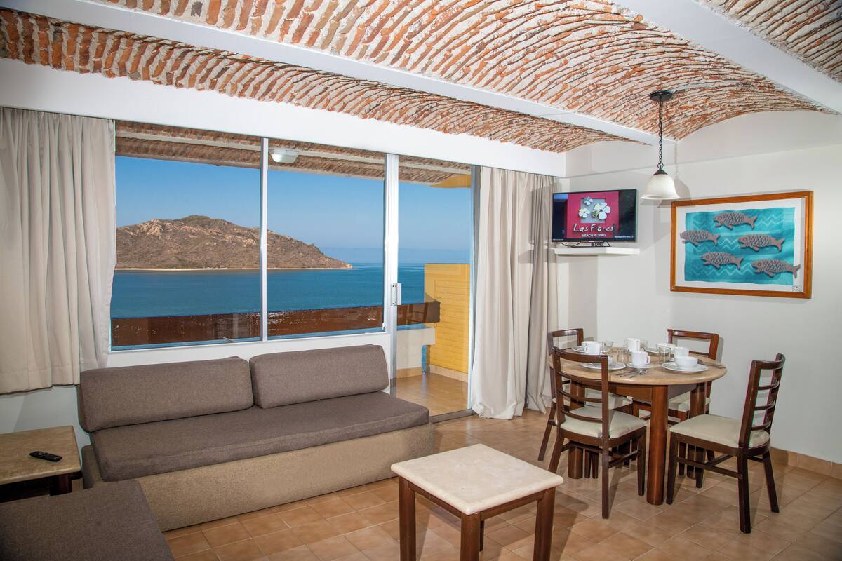 Ein Zimmer mit einer Couch, einem Esstisch mit Stühlen, einem Fernseher und Meerblick.