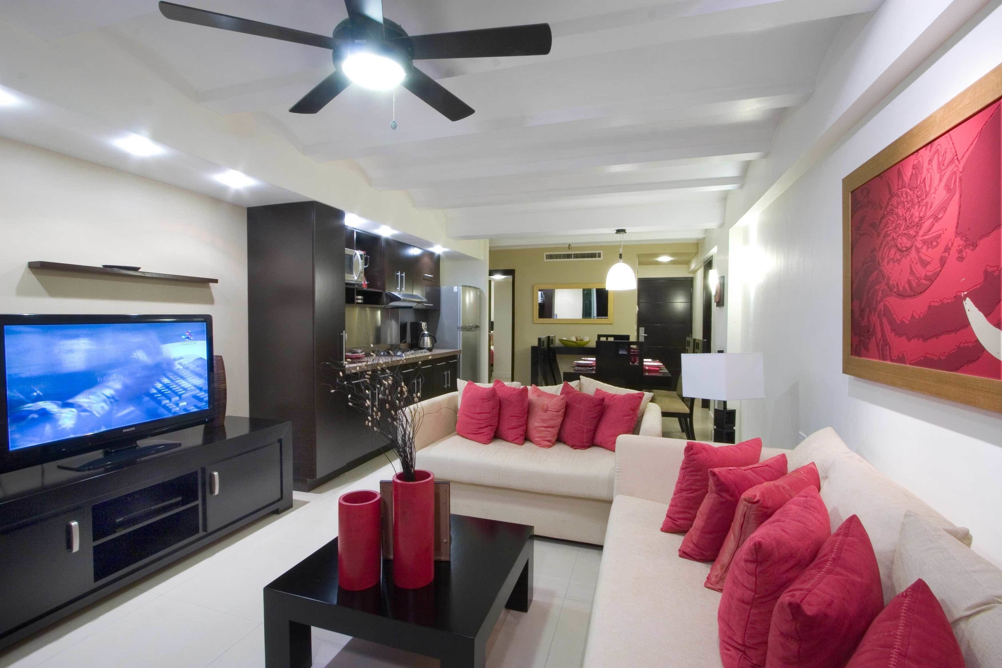 A modern living room with a flat-screen TV, a black coffee table, a beige sofa with red cushions, and a ceiling fan.