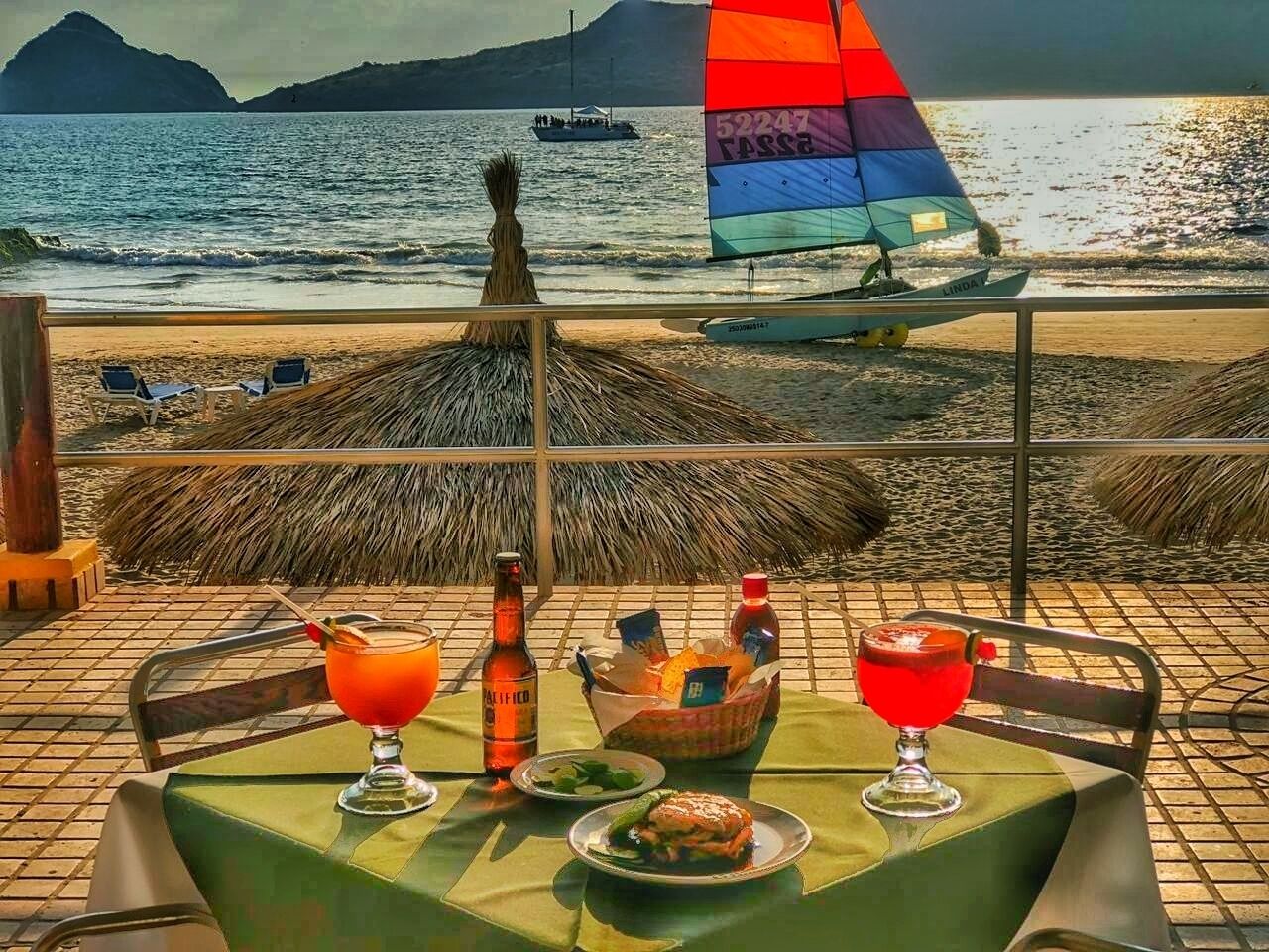 Una mesa con bebidas y aperitivos, una botella y una cesta en una terraza con vista al mar y un velero.