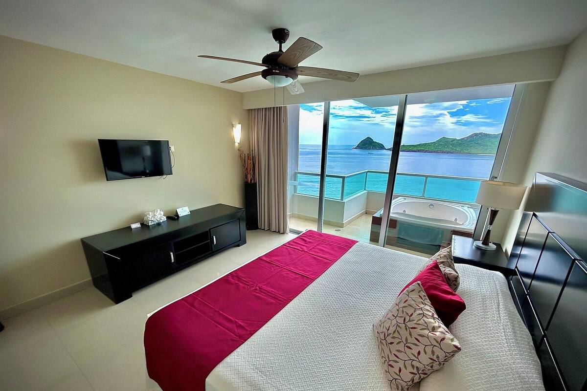 Ein Hotelzimmer mit Bett, Fernseher, Deckenventilator, Balkon mit Meerblick und einem kleinen Tisch mit Lampe.