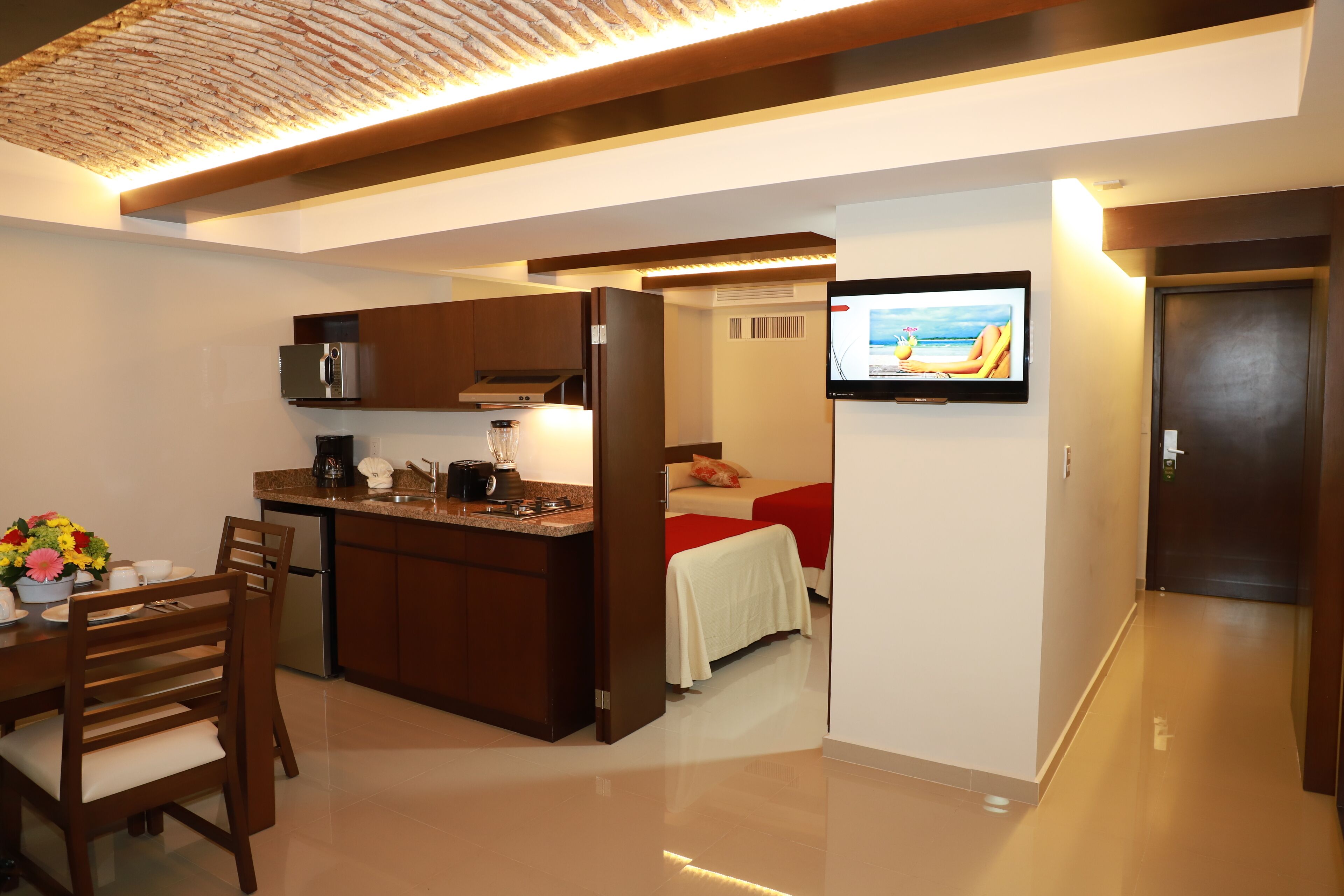 A compact hotel room with a kitchenette, a dining area, and a TV mounted on the wall.