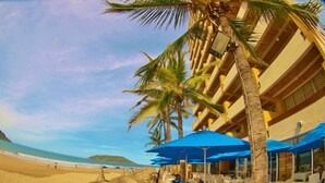 Un hotel frente a la playa con sombrillas de paja, sillones blancos y un comedor con mesas y sillas.