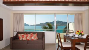 Una sala de estar con sofá, una mesa de comedor para dos personas y un ventanal con vistas al mar y a las montañas.