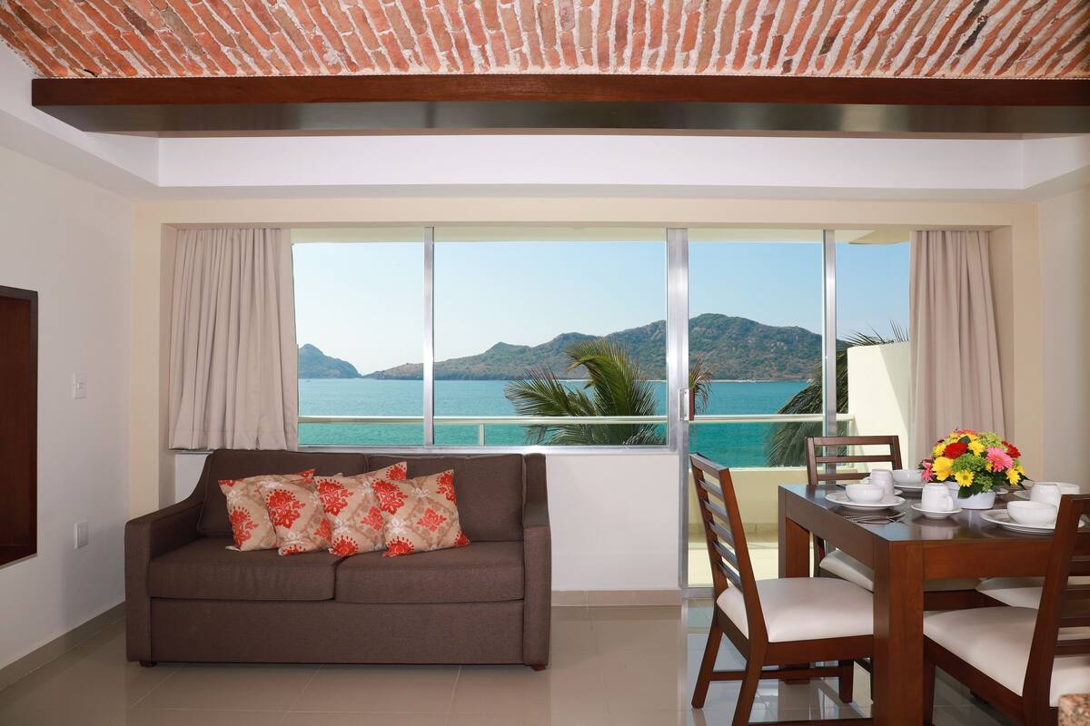 Ein Wohnzimmer mit einer Couch, einem Esstisch für zwei Personen und einem großen Fenster mit Blick auf das Meer und die Berge.