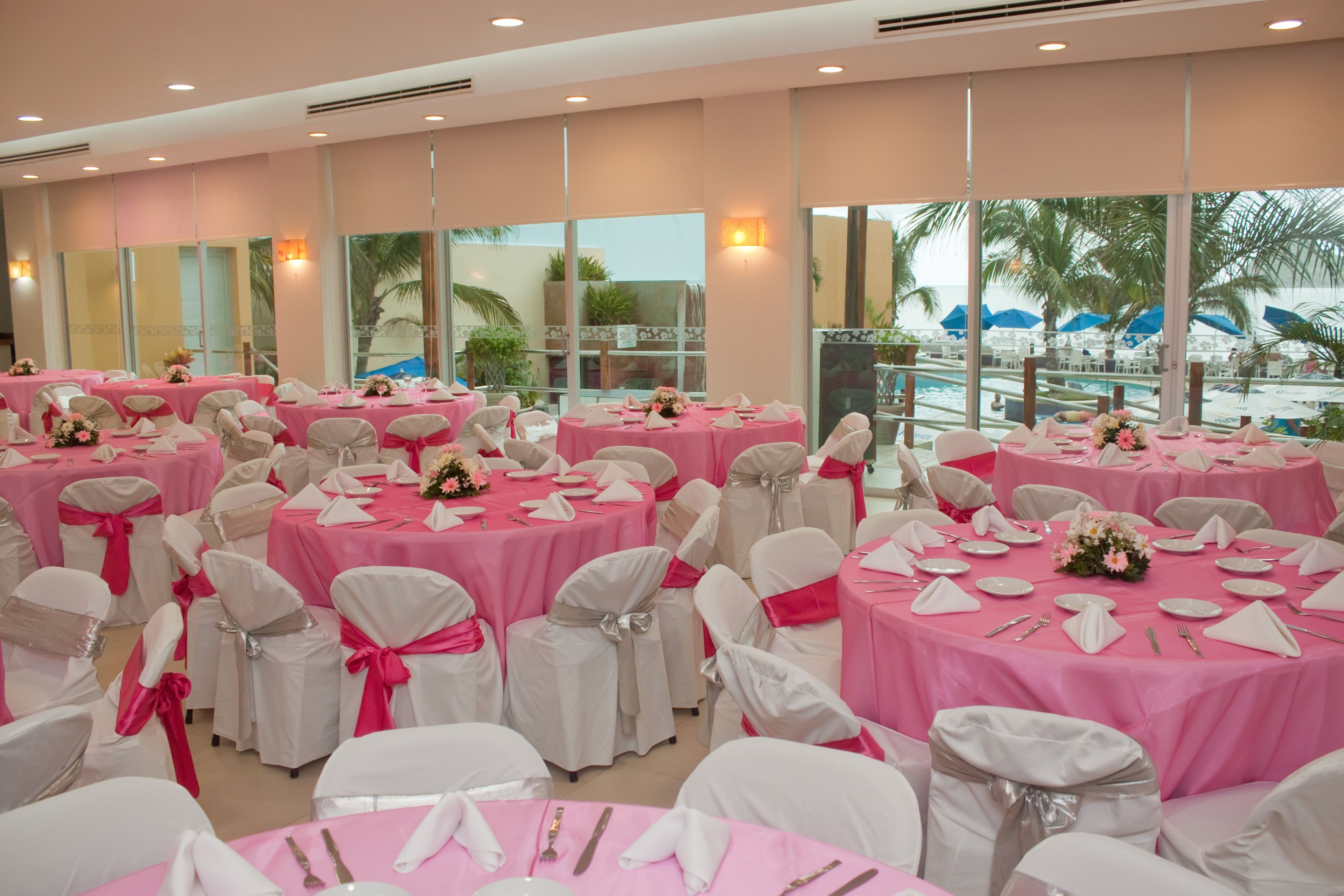 A banquet hall with pink tables and white chairs, large windows, and a view of a pool area.