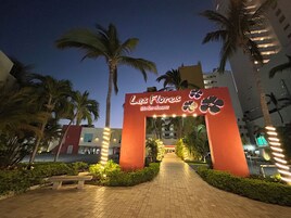 Una entrada al Las Flores Beach Resort con un arco llamativo, palmeras y un camino pavimentado que conduce al edificio.