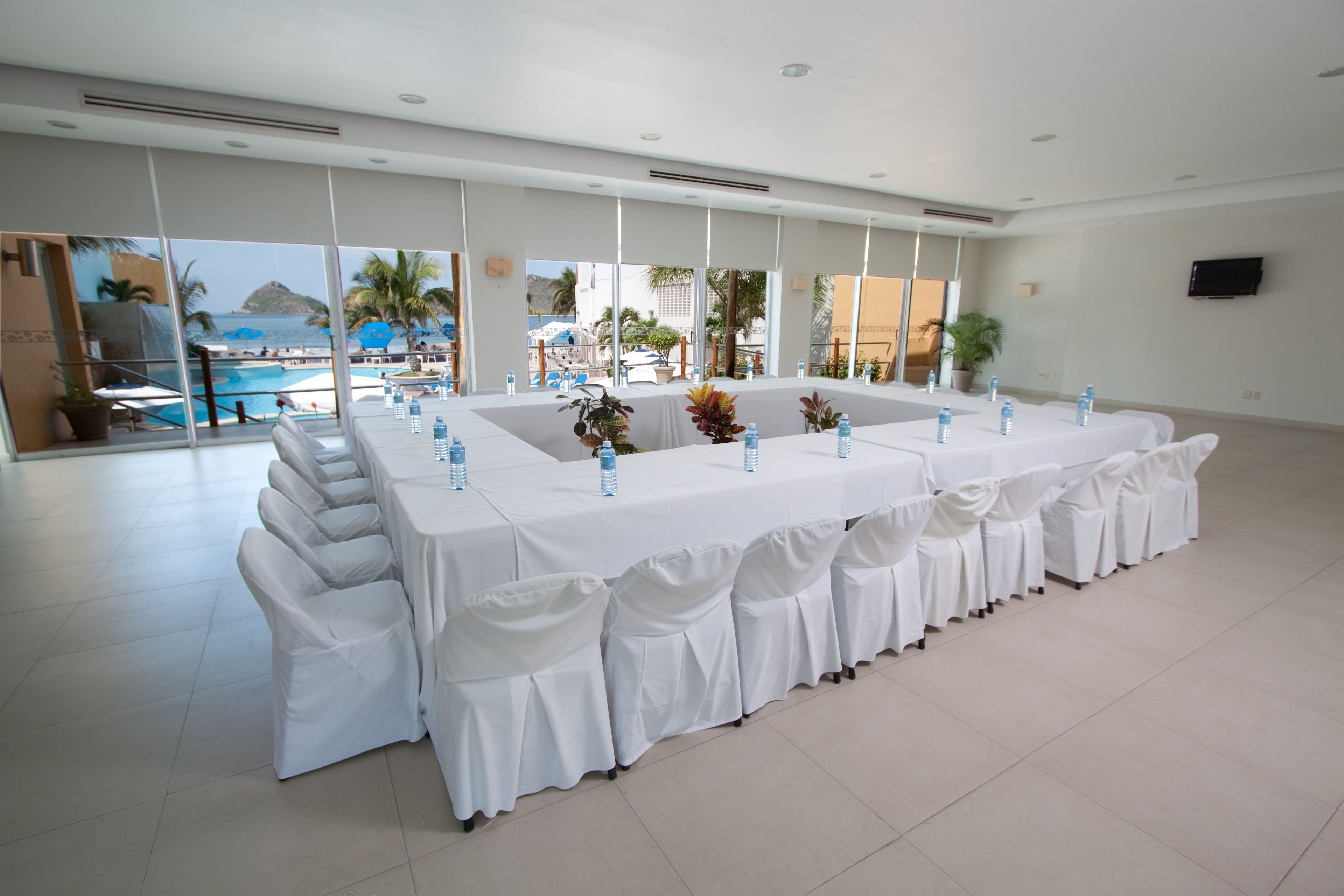 A large, well-lit room with a long table set for a meeting, large windows with a view of a pool and palm trees, and a flat-screen TV on the wall.