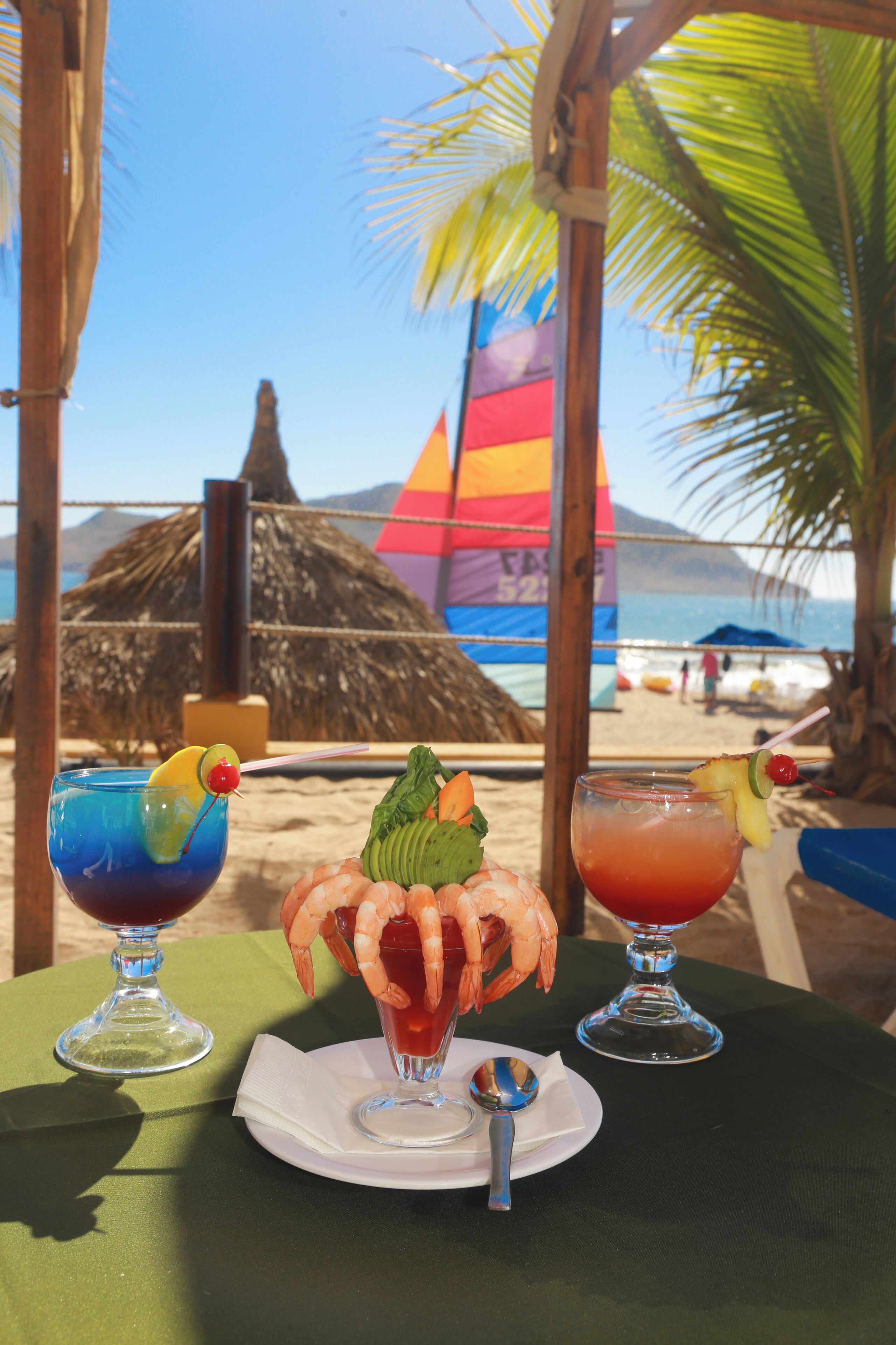 A table with two drinks and a shrimp cocktail, set against a beach backdrop with a thatched structure and a colorful flag.