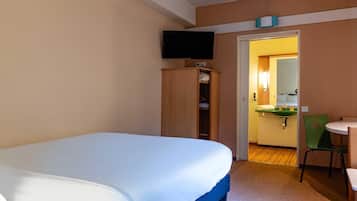 A hotel room with a bed, a television mounted above it, a wardrobe, a small kitchenette, and a chair.