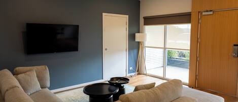 Un salon moderne avec un canapé d’angle, un téléviseur à écran plat fixé au mur, une petite table basse noire et une porte-fenêtre coulissante donnant sur un balcon.