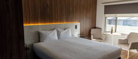 A modern hotel room with a large bed, a wooden accent wall, a small round table, and a window with a view.