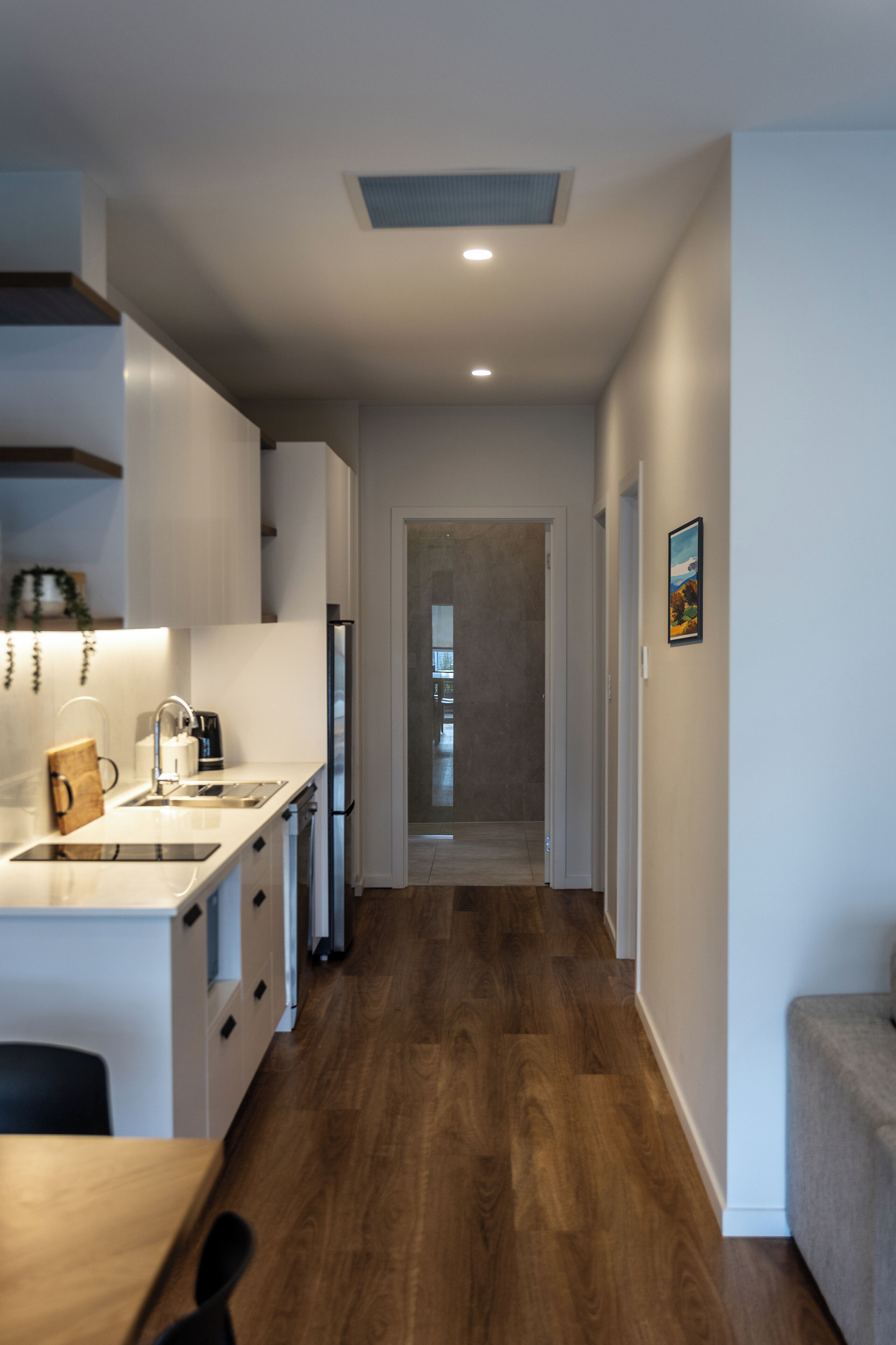 A modern kitchen with white cabinetry, a wooden countertop, and a built-in oven. A hallway with wooden flooring leads to a living area.