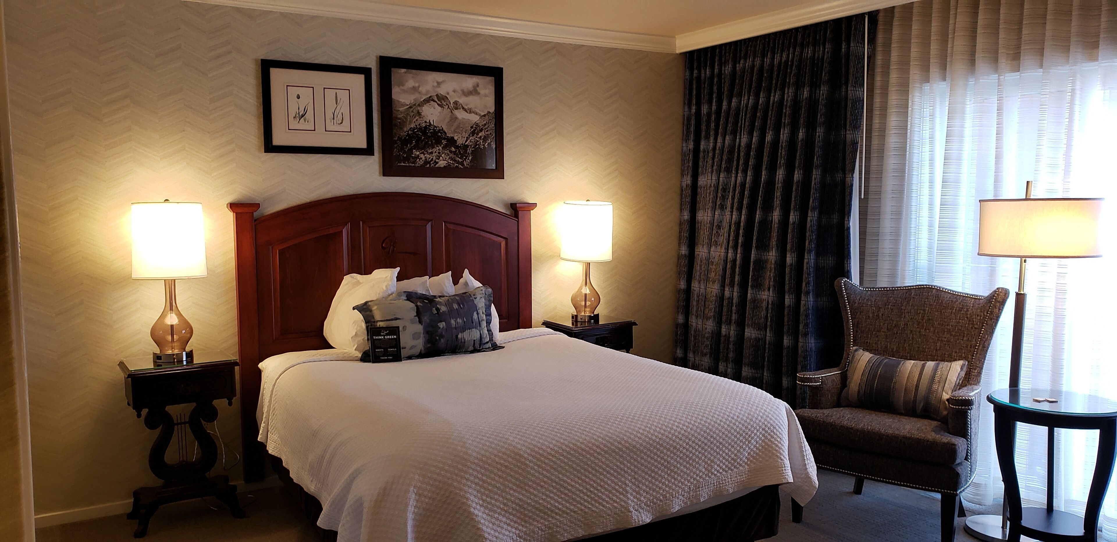 A hotel room with a large bed, two bedside lamps, a chair, and a table. There are two framed pictures on the wall.