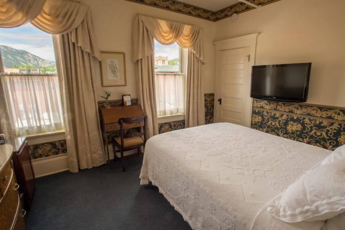 A bedroom with a bed, a desk, a chair, a TV, and a window with curtains.