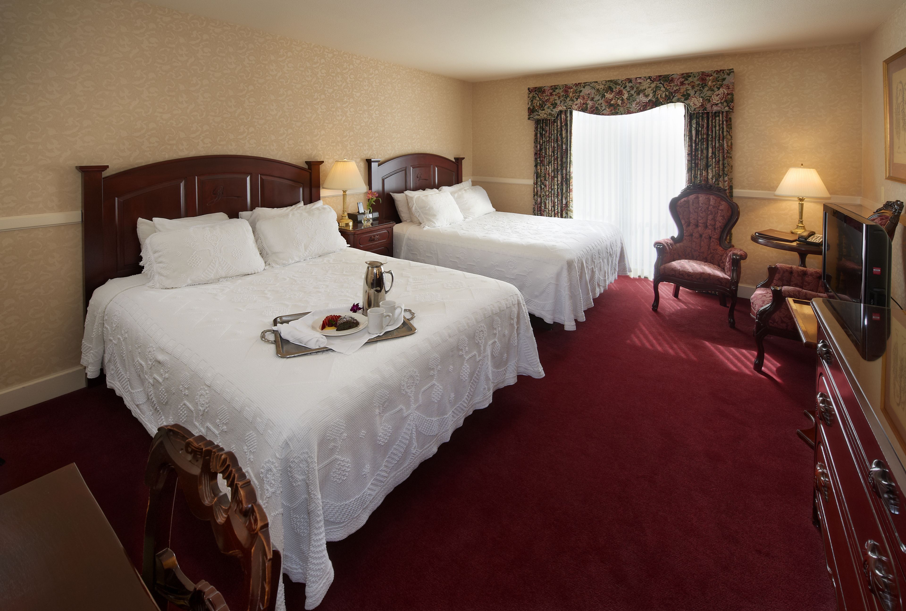 A hotel room with two beds, a red carpet, a wooden chair, a television, and a small table with a lamp.