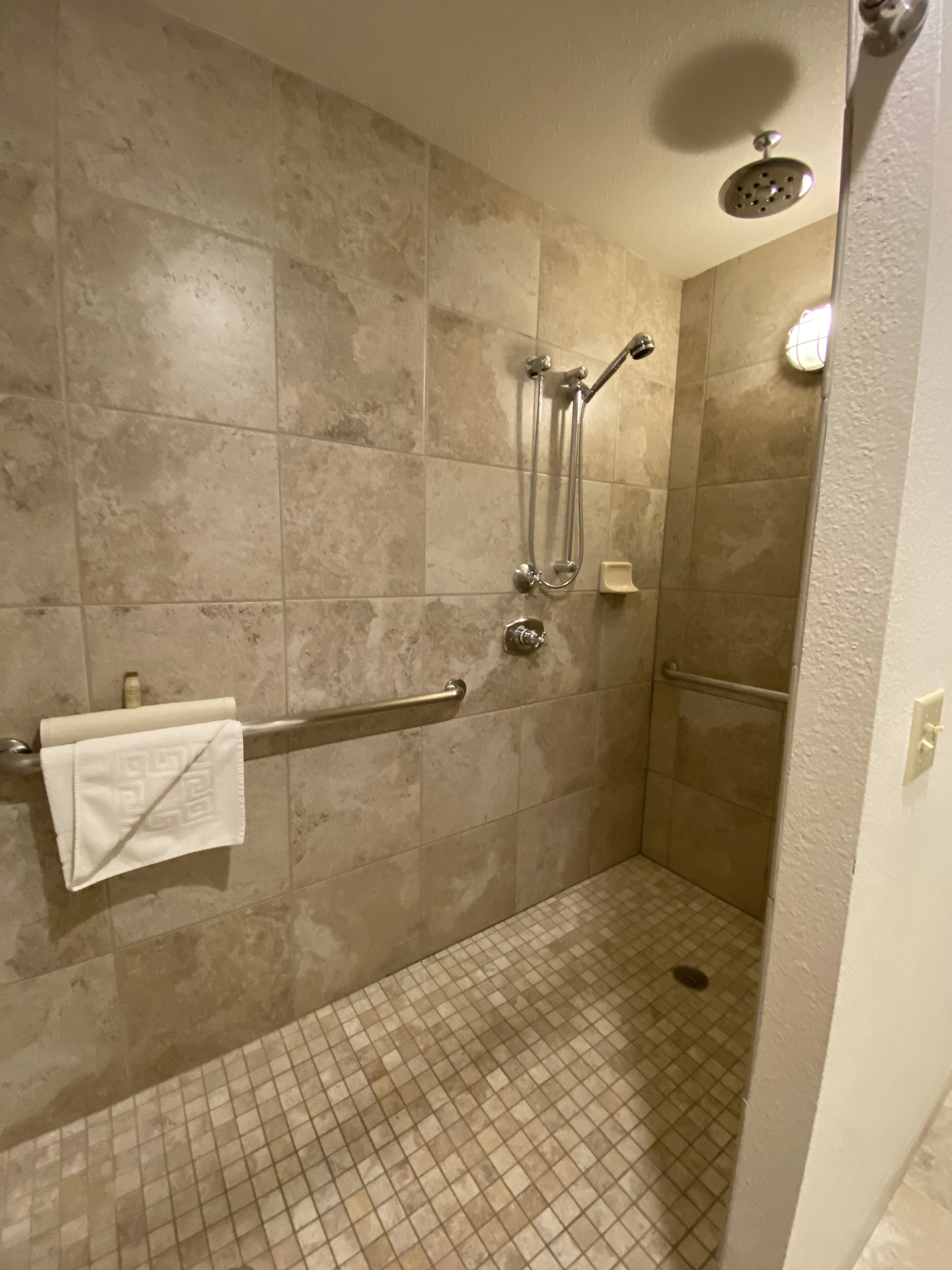A shower area with beige tiles, a handrail, a towel, and a handheld showerhead.