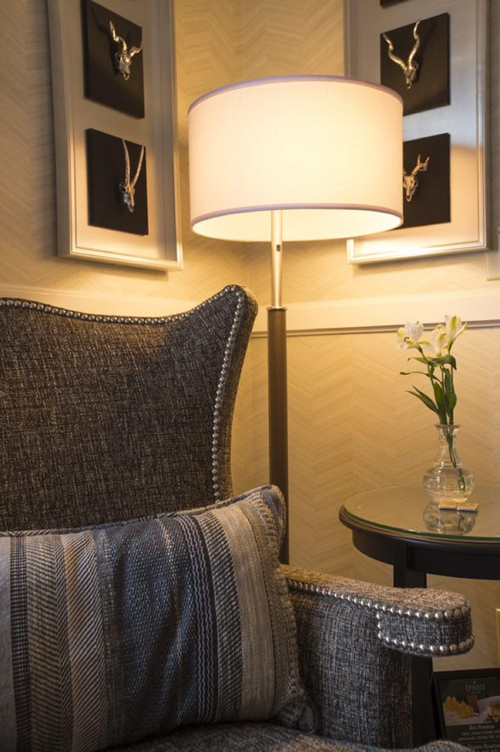 Una habitación con un sillón estampado, una lámpara de pie, un jarrón con flores y cuadros en la pared.