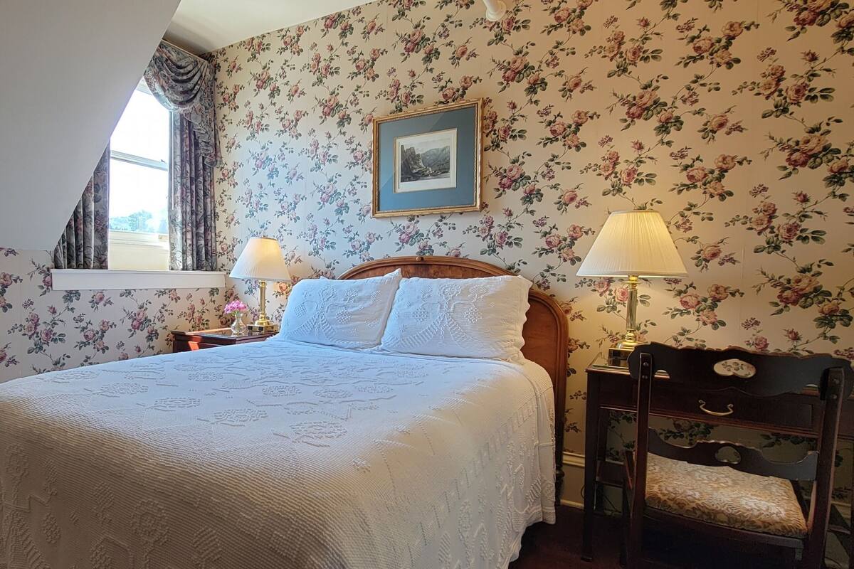 A bedroom with floral wallpaper, a bed with white linens, two bedside lamps, a wooden nightstand, a chair, and a framed picture on the wall.