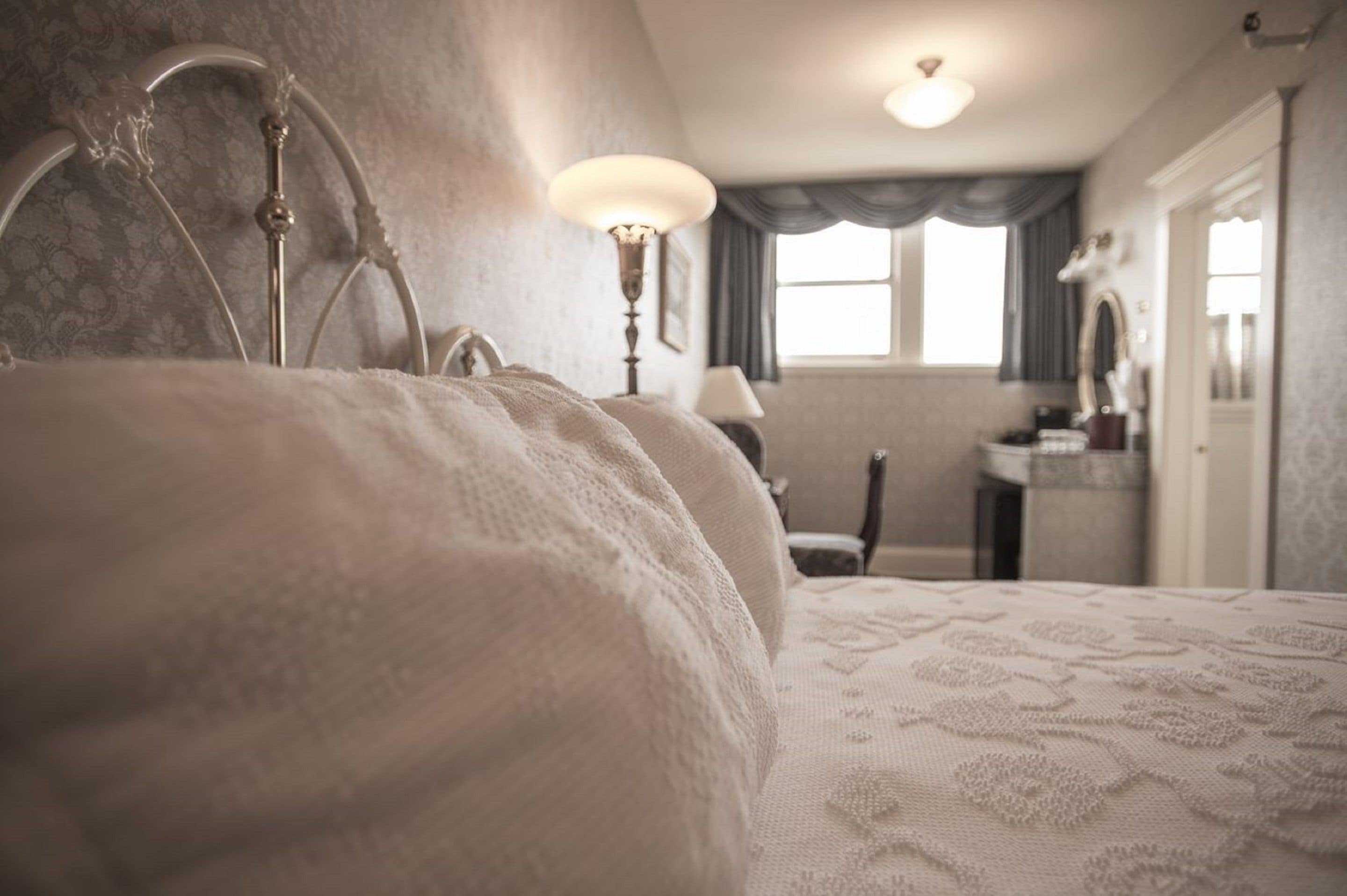 A bedroom with a large bed, a bedside table, a lamp, a mirror, and a window with curtains.
