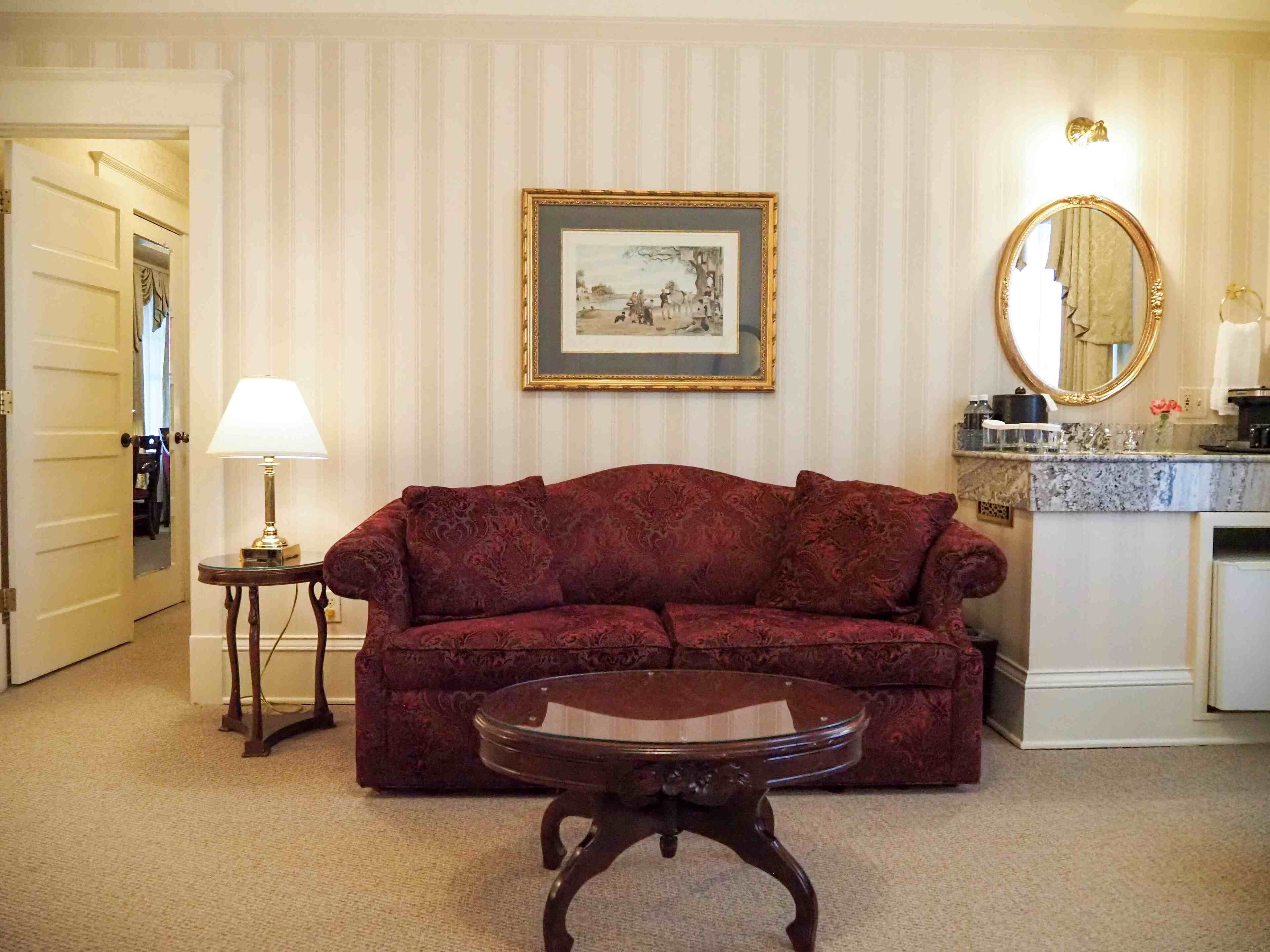 A living room with a red sofa, a glass coffee table, a framed picture on the wall, a lamp on a side table, and a kitchen area with a marble countertop and a sink.