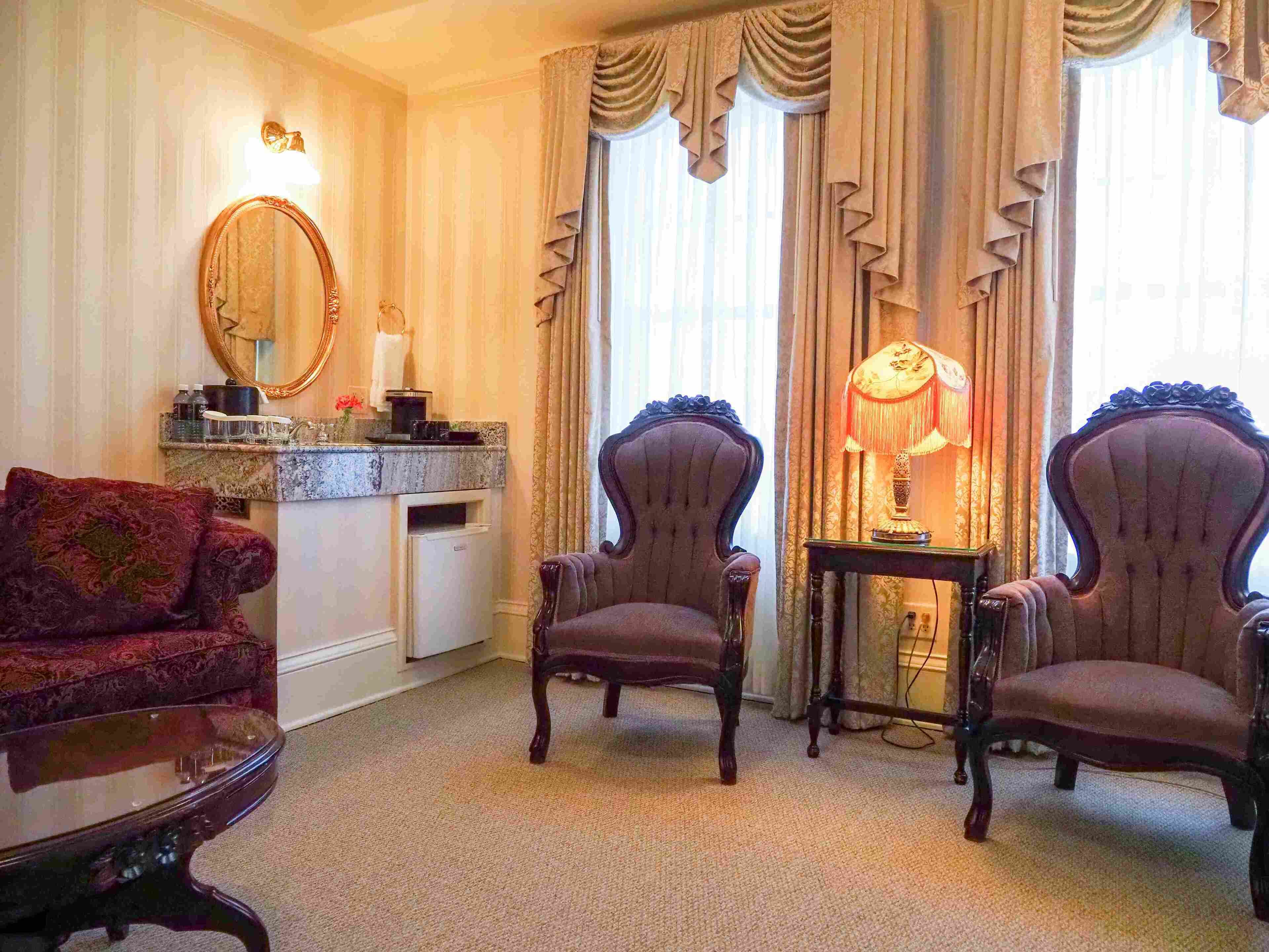 Una habitación de estilo vintage con dos sillones, un sofá, una mesita con una lámpara, un espejo y un tocador con lavabo.