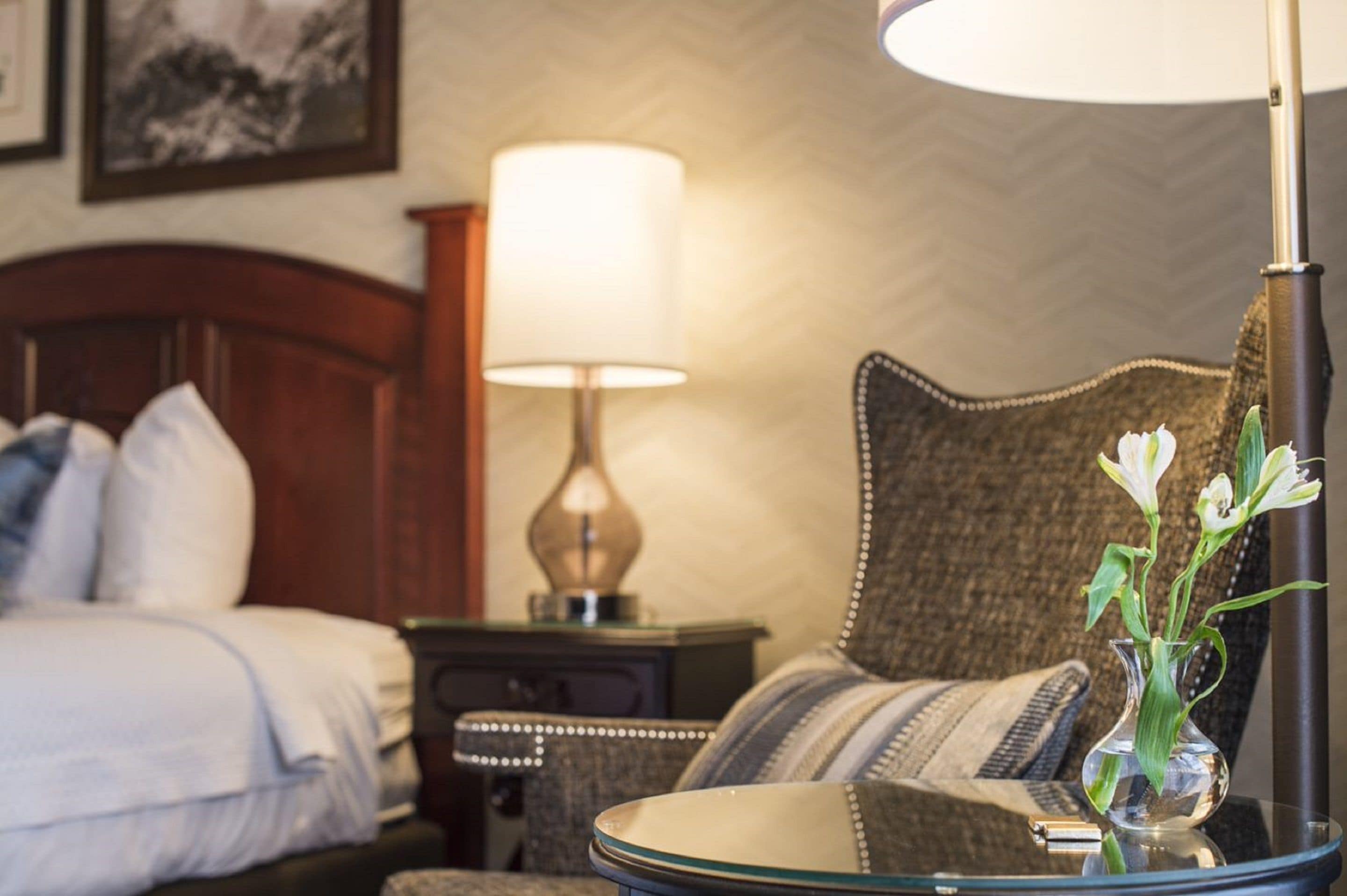 A hotel room with a bed, a nightstand with a lamp, a chair, and a vase with flowers on a table.