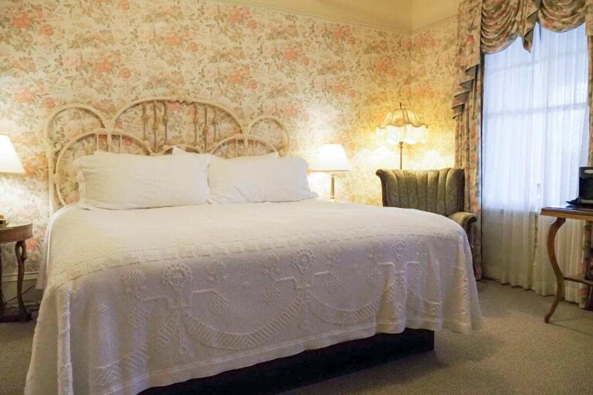A bed with a white bedspread, a wooden headboard, two bedside lamps, a chair, and a television on a wooden table.