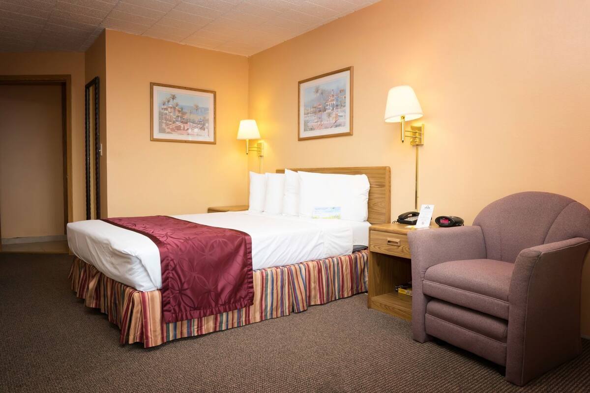 A hotel room with a bed, a chair, two wall-mounted lamps, and two framed pictures on the wall.