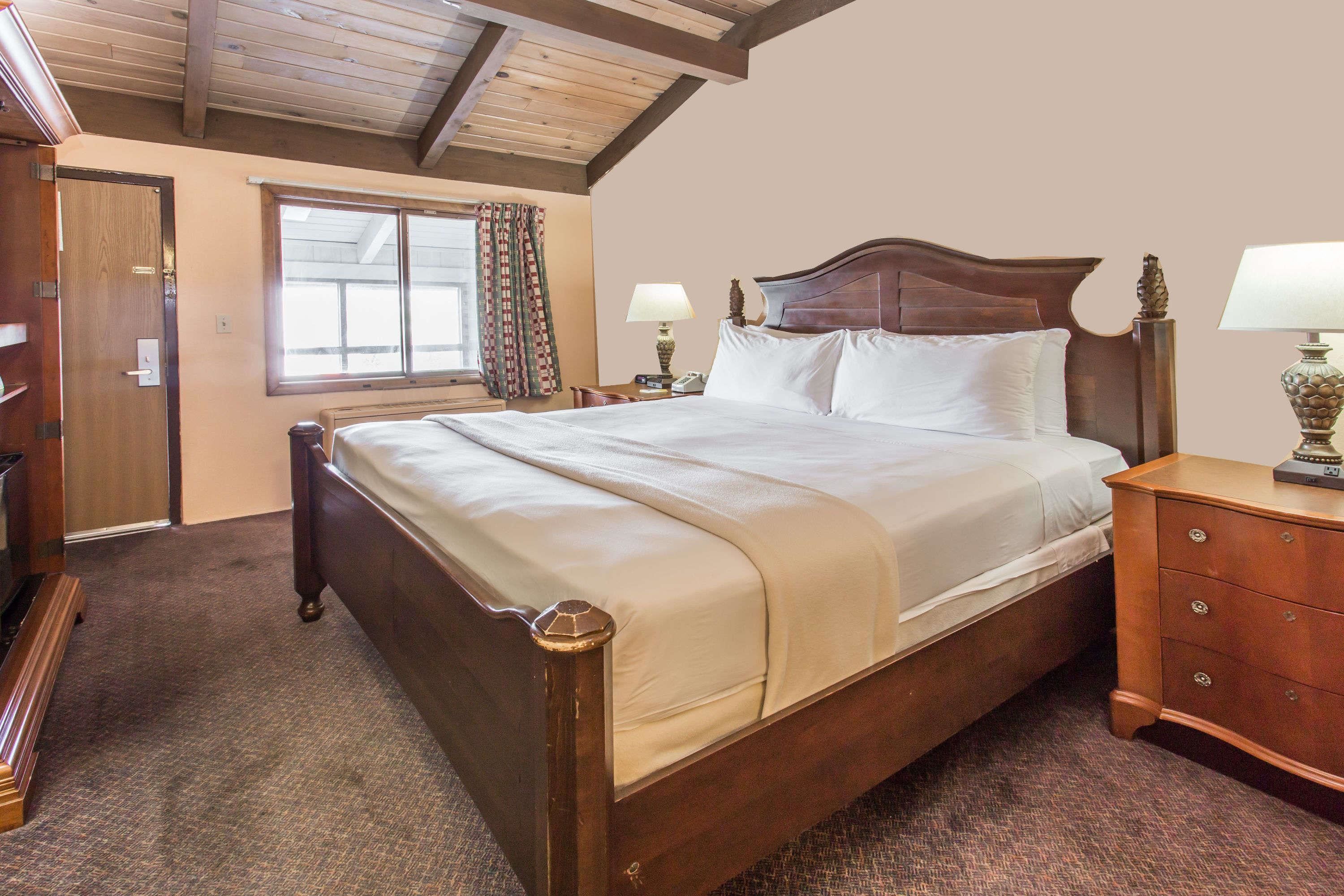 Uma cama de madeira com dossel e roupa de cama branca, um criado-mudo de madeira com uma luminária, um aparador de madeira e uma janela com cortinas.