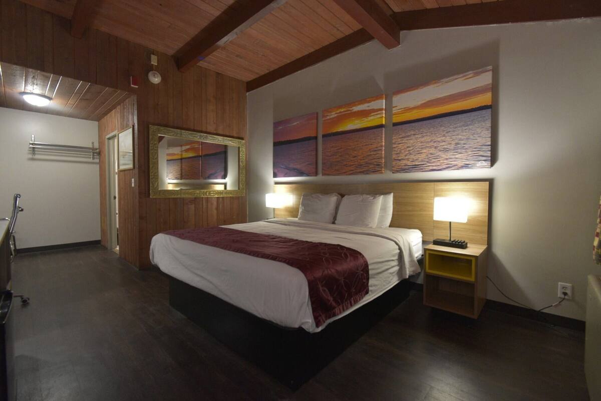 A hotel room with a large bed, wooden ceiling beams, a painting of a sunset, and a bedside table with a lamp.